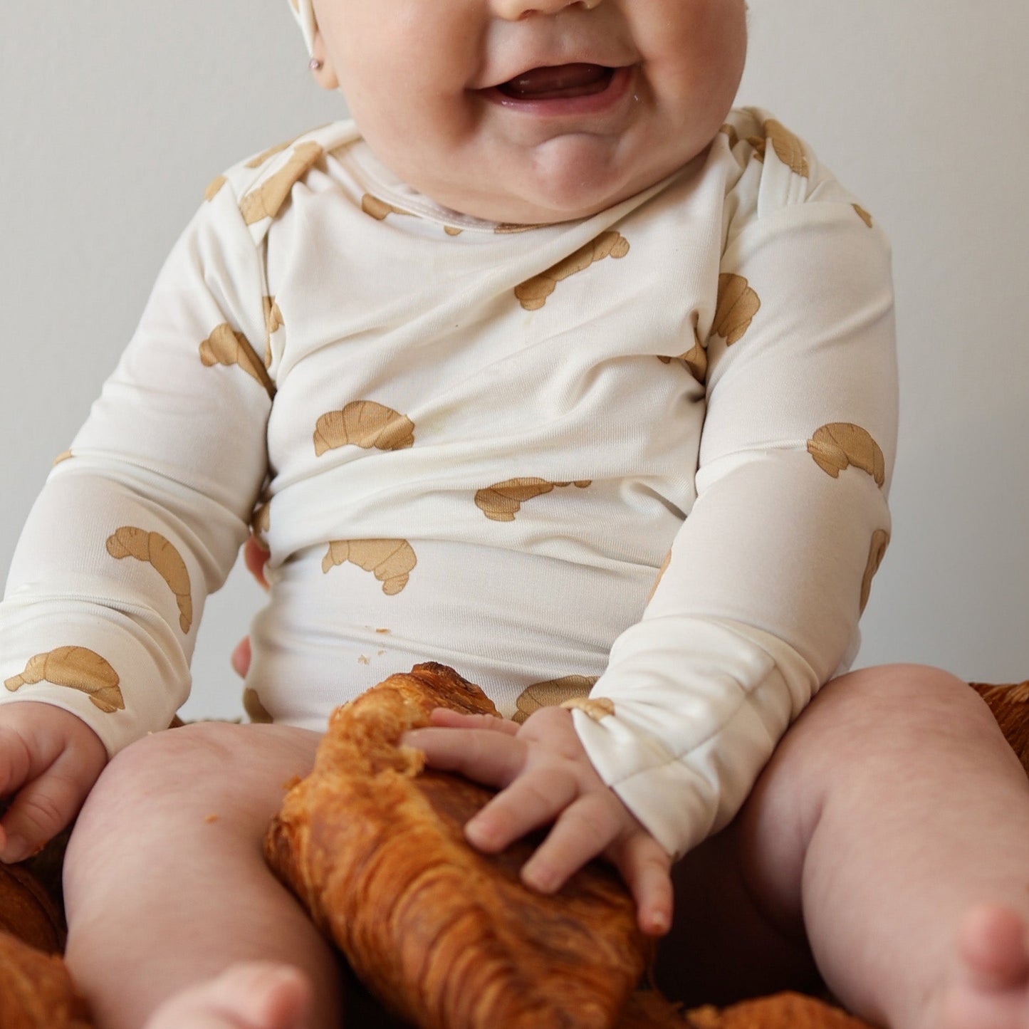 A smiling baby in the forever french baby Long Sleeve Bamboo Snapsuit | Croissants sits among large croissants, reaching out to touch one.