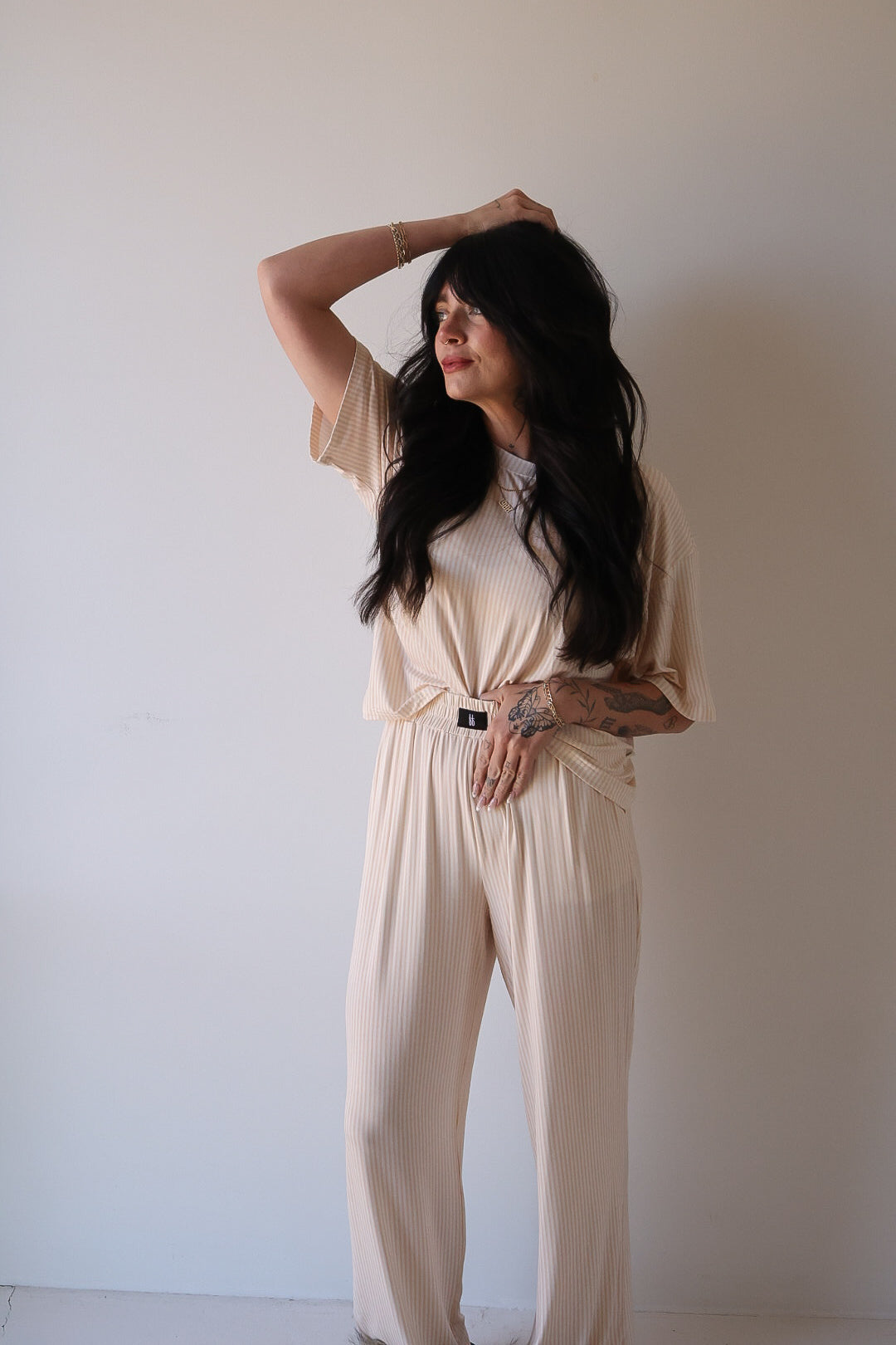 A woman with long dark hair stands against a plain light wall, wearing the Women's Boxy Tee Lounge Set in Sadie Stripe by forever french baby. She touches her head and gazes to the side, radiating a relaxed vibe.