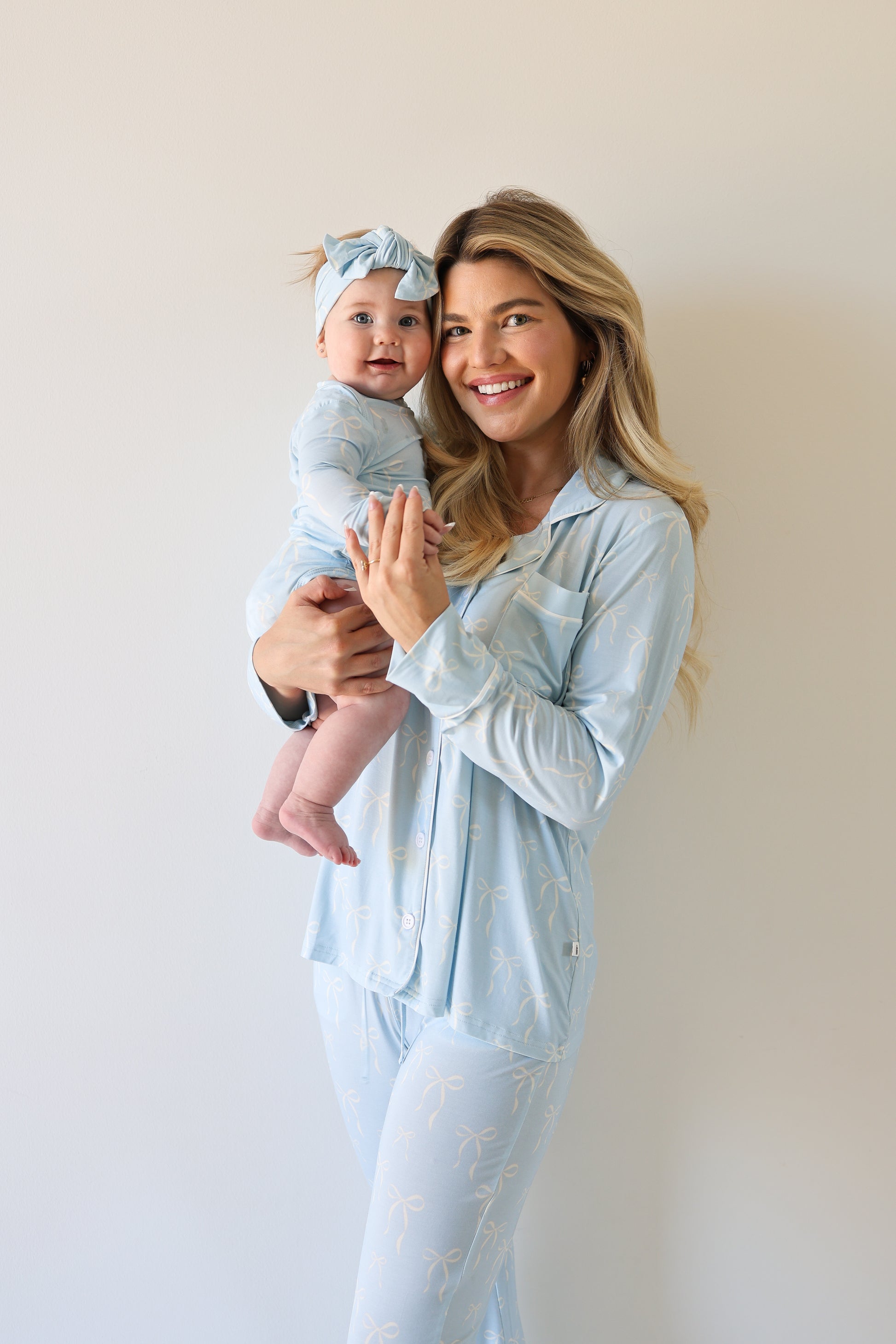A smiling woman in the forever french baby Women's Bamboo Button Up Lounge Set | Blue Bows holds a baby in matching blue pajamas and a headband, both standing against a plain, light-colored background.