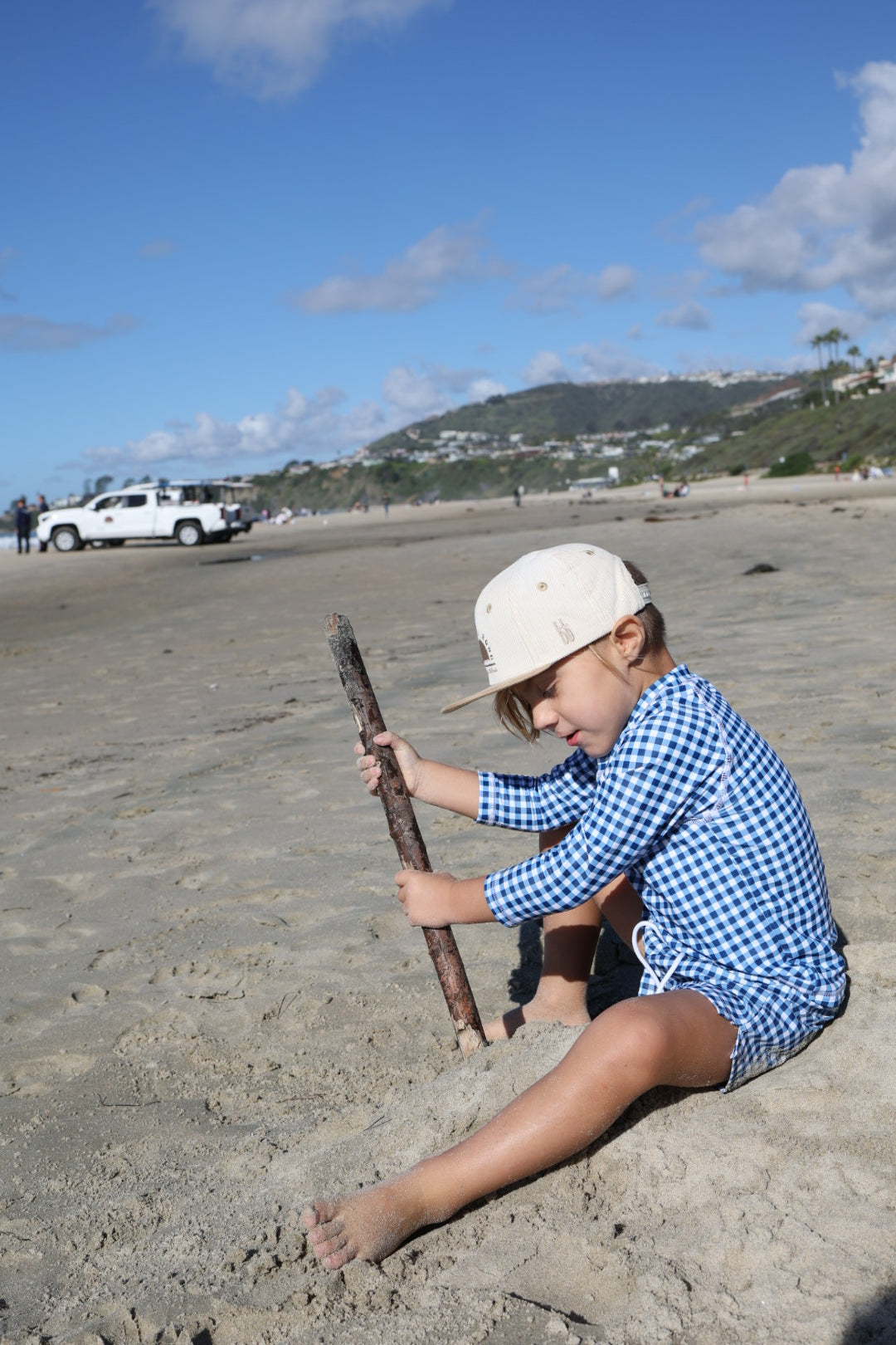 A young child wearing forever french baby’s Child Board Short in Blue Gingham and a beige cap sits on the sandy beach, digging with sticks. Blue sky, distant hills, and parked cars appear in the background.
