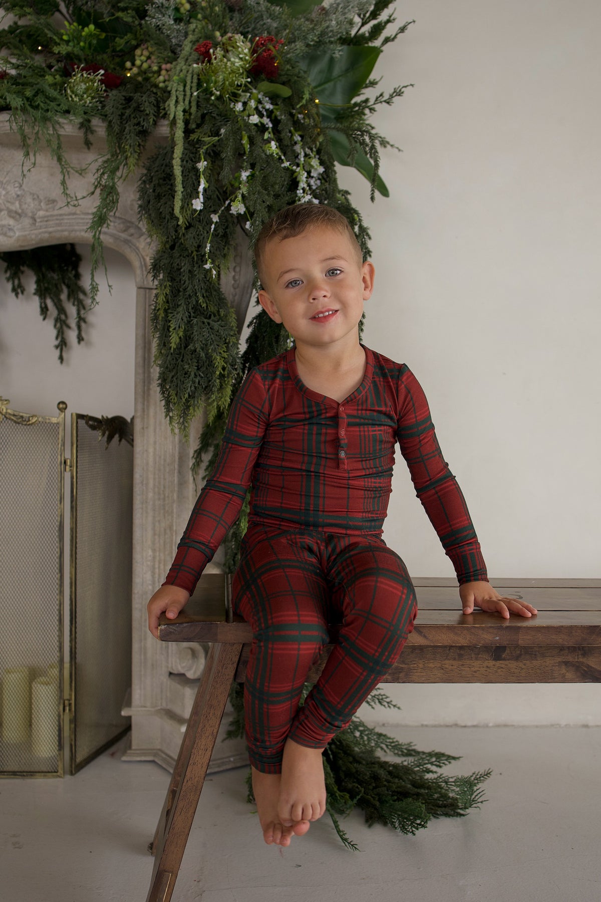 A young boy sits on a wooden bench wearing a long-sleeved Bamboo Two Piece Pajamas | Christmas Plaid set with a red and green plaid pattern.