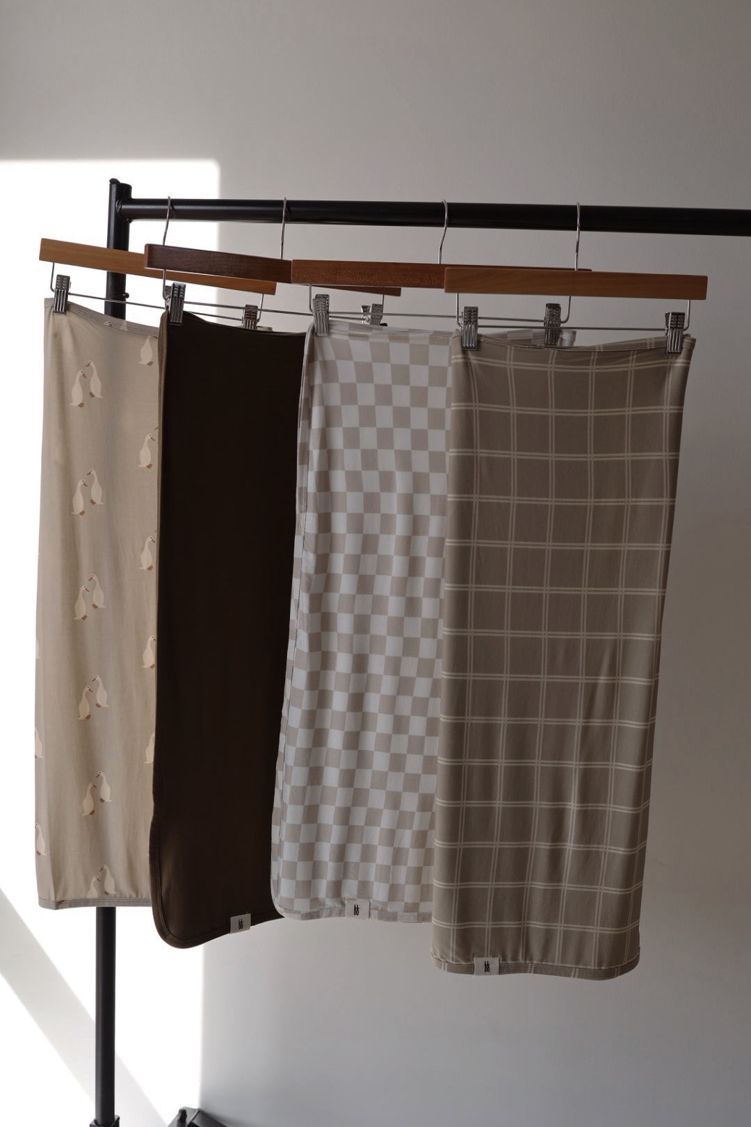 Four Bamboo Swaddles from forever french baby’s Silly Goose collection, in beige and brown with abstract, solid, checkerboard, and grid patterns, hang on wooden hangers from a black rack, softly illuminated by sunlight.