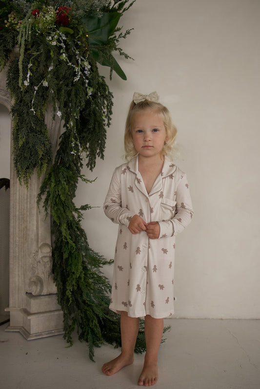 A young child wearing a breathable, tan, button-down Bamboo Sleeping Dress | Gingersnap featuring a brown gingerbread man print.