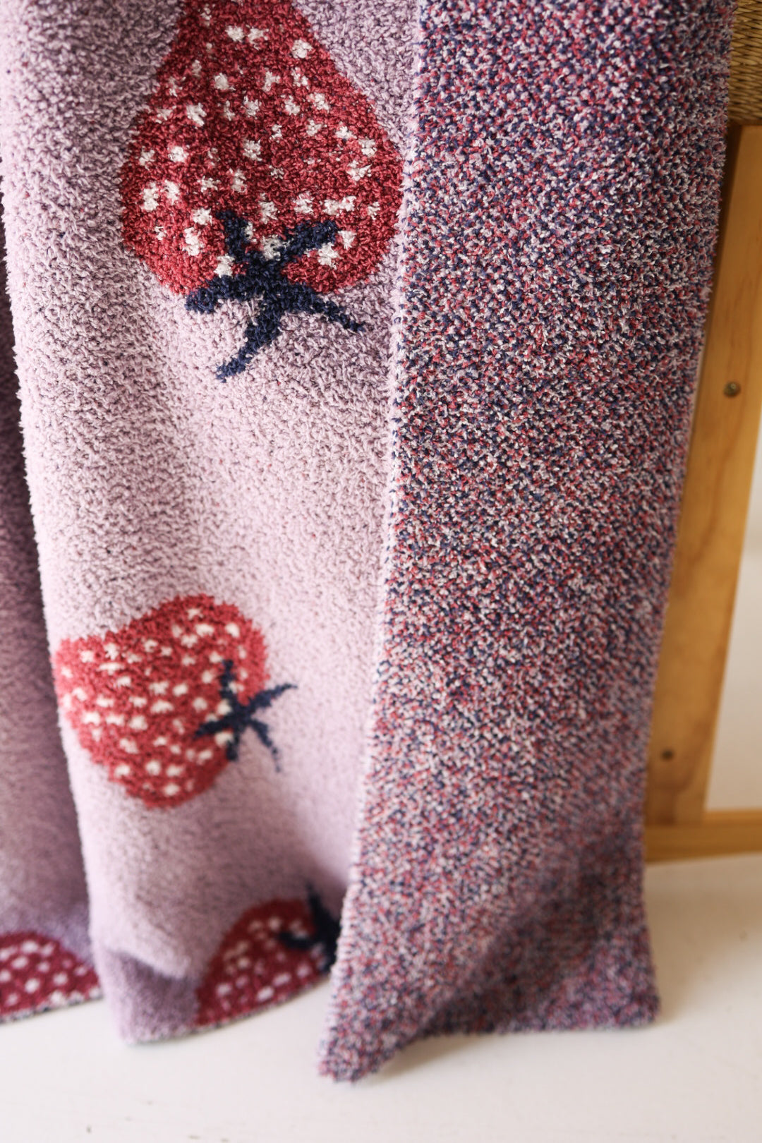 A close-up of the forever french baby Plush Blanket | Matilda, an OEKO-TEX certified microfiber blanket with large red strawberries and green stems on light purple, plus a speckled purple and red section, draped over a wooden chair.