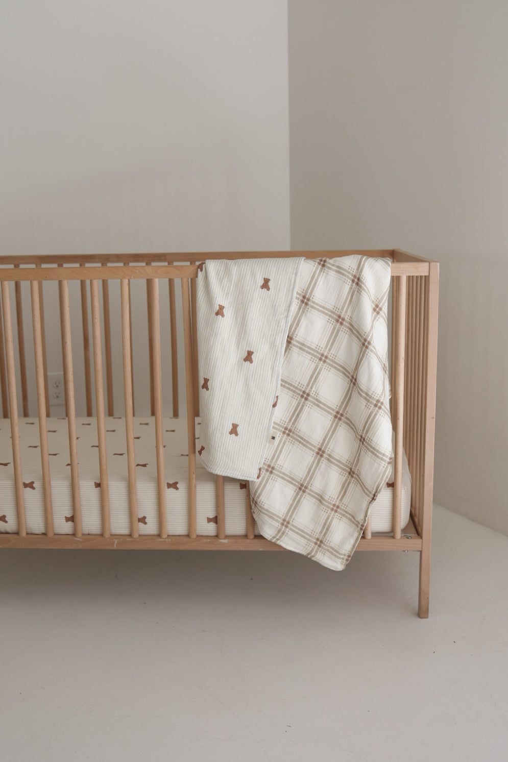 A forever french baby Muslin Crib Sheet | Teddy covers the wooden crib in a minimalist room, with two cozy blankets—one featuring brown bear prints and the other a beige plaid—draped over the rail for stylish baby bedding.