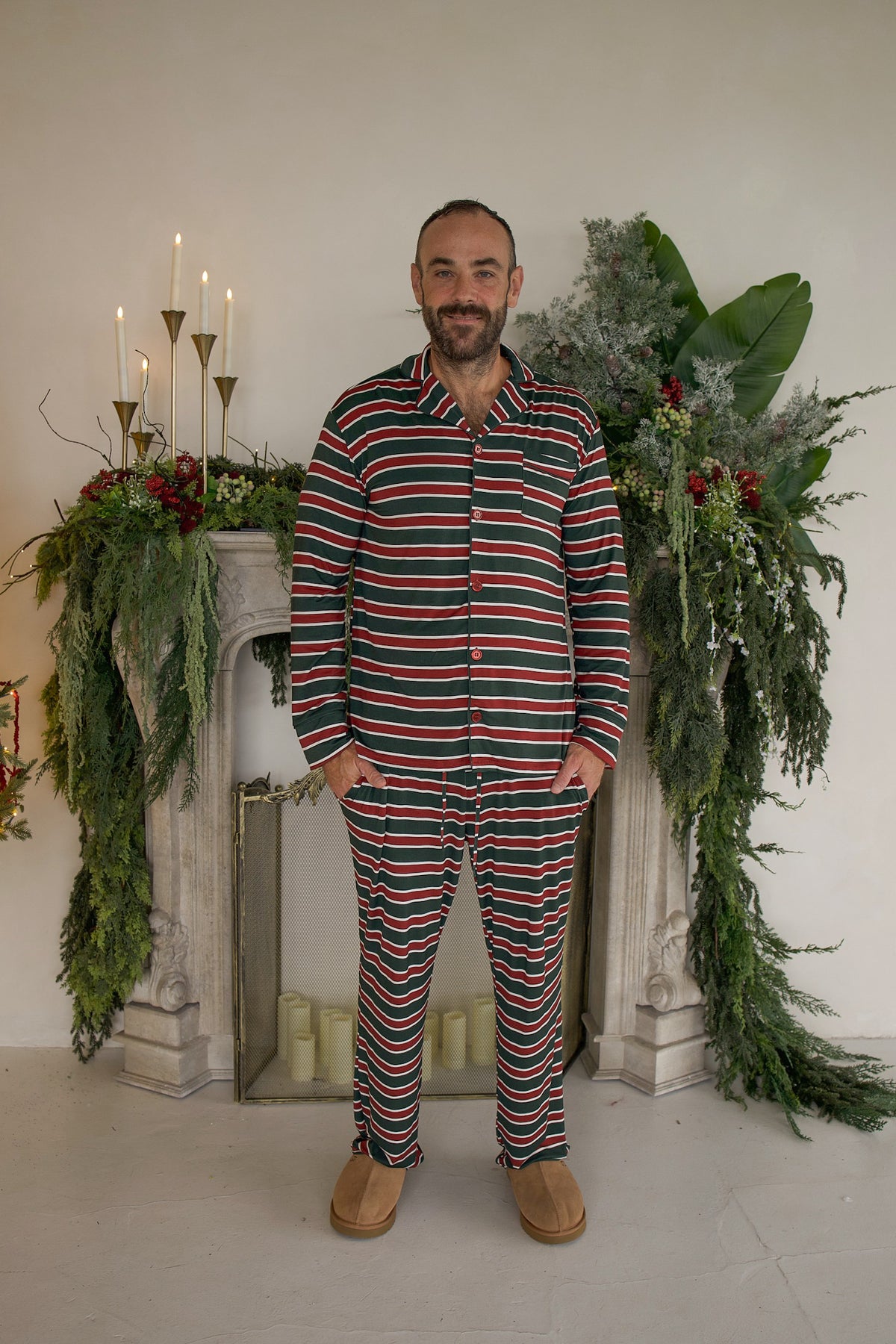A man wearing a Men’s Bamboo Button Up Lounge Set | Holiday Stripe featuring red, green, and white stripes, standing in front of a decorative fireplace.