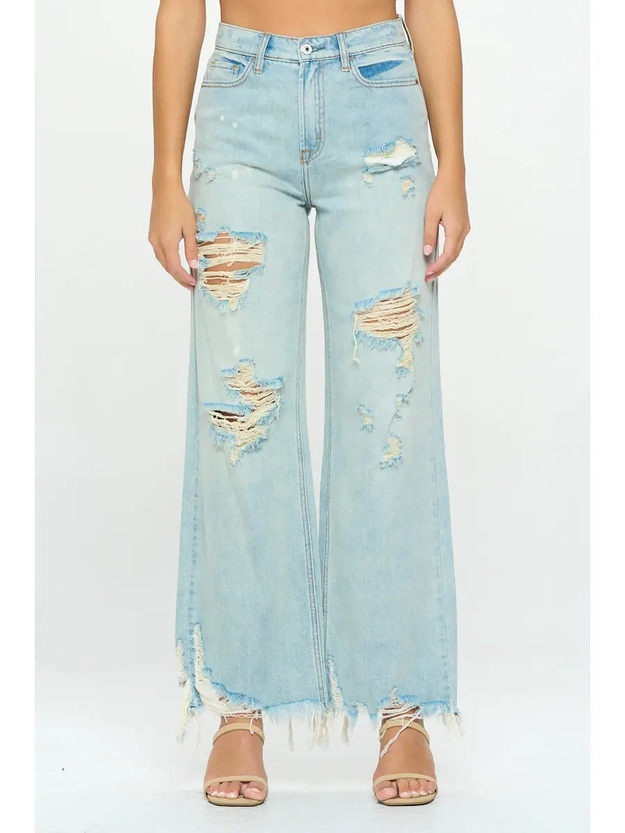 A person is wearing I&M JEAN, INC. High Rise Distressed Skater Jeans in a trendy light wash, featuring a high-waisted fit with a frayed hem and multiple rips and tears. They pair these jeans with nude strappy heeled sandals against a plain white background.