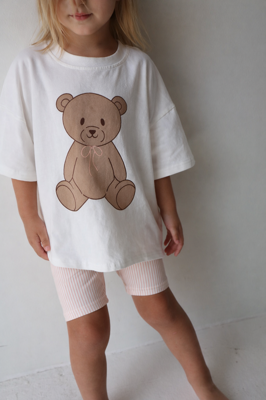 A young child wears the forever french baby Child Short Set | Lottie, featuring a teddy bear graphic tee and light pink and white bike shorts, posed against a plain background with her face partially visible.