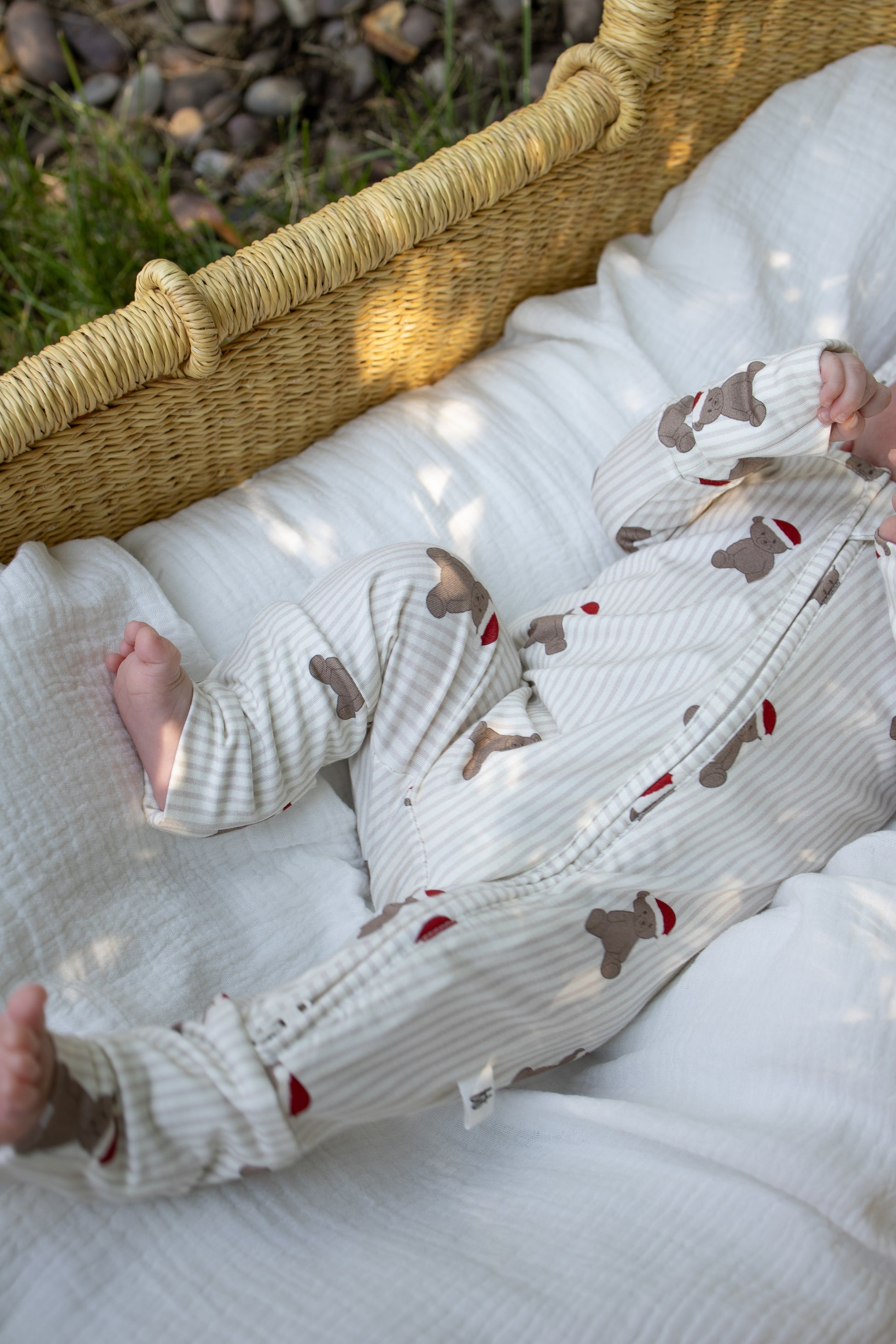 A baby wearing forever french baby’s Bamboo Zip Pajamas in the Santa Teddy print lies in a woven basket lined with a white blanket, basking in gentle outdoor sunlight—perfect for highlighting our hypoallergenic, cozy, and comfortable bamboo baby clothing.