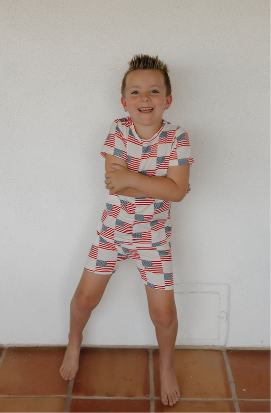 A young boy stands barefoot on a tiled floor, smiling with arms crossed. He wears the forever french baby Bamboo Short Set | Americana, a breathable sleepwear set decorated with American flags for all-night comfort.