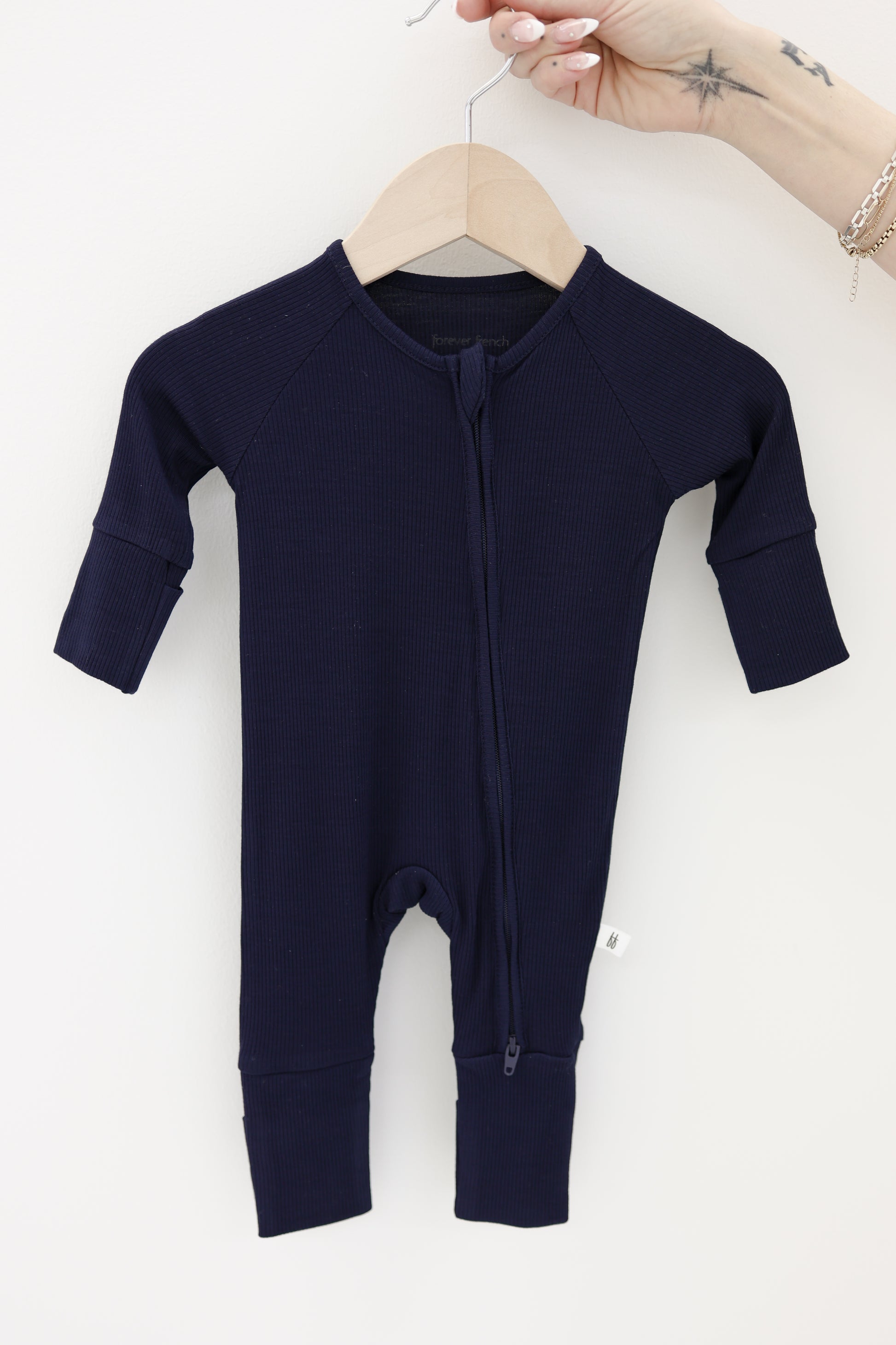 A hand holds a hanger displaying the forever french baby Bamboo Zip Pajamas | Blueberry Ribbed, a long-sleeved, navy blue baby onesie with a front zipper, set against a plain white background.
