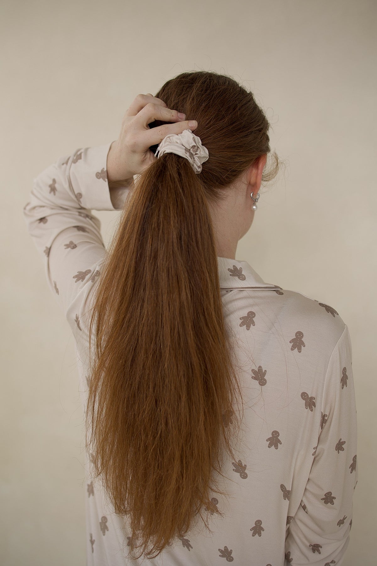 A person with long hair wearing a light-colored Gingersnap Scrunchie by forever french baby, decorated with a print of small gingerbread men.