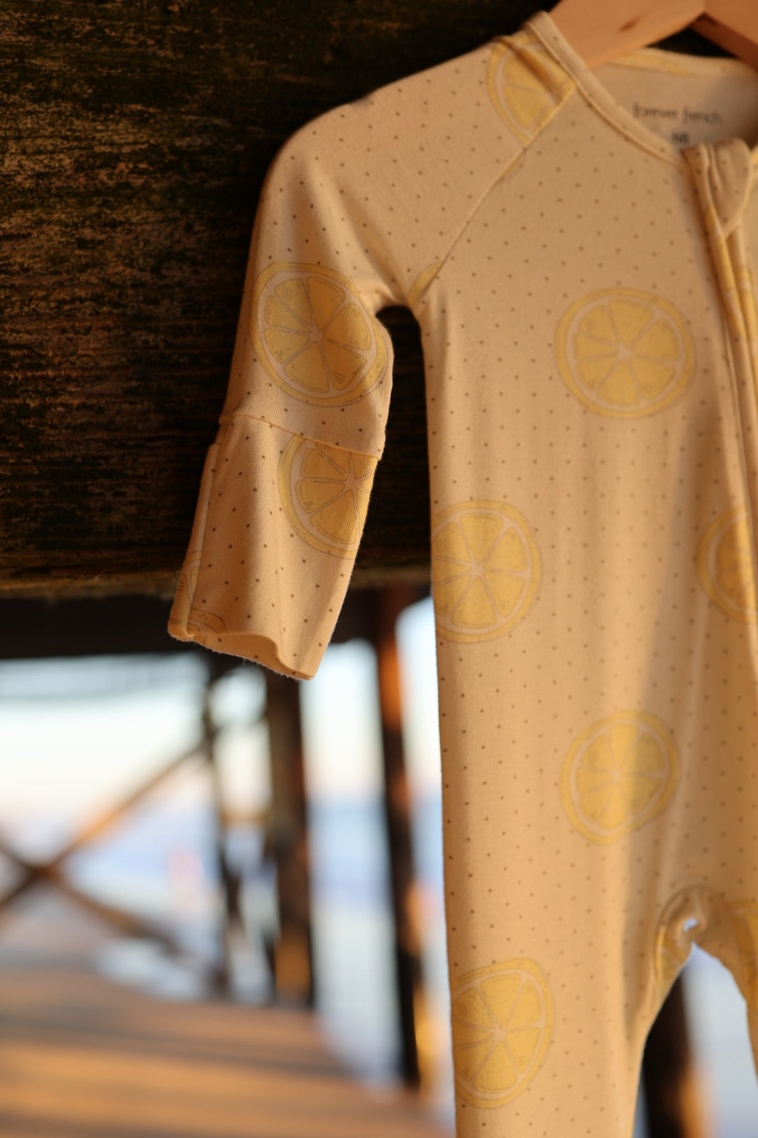 The Bamboo Zip Pajamas | Lemon Drop by forever french baby, featuring yellow citrus slices and small dots on beige breathable fabric, hangs on a wooden structure with sunlight and beams softly blurred in the background.
