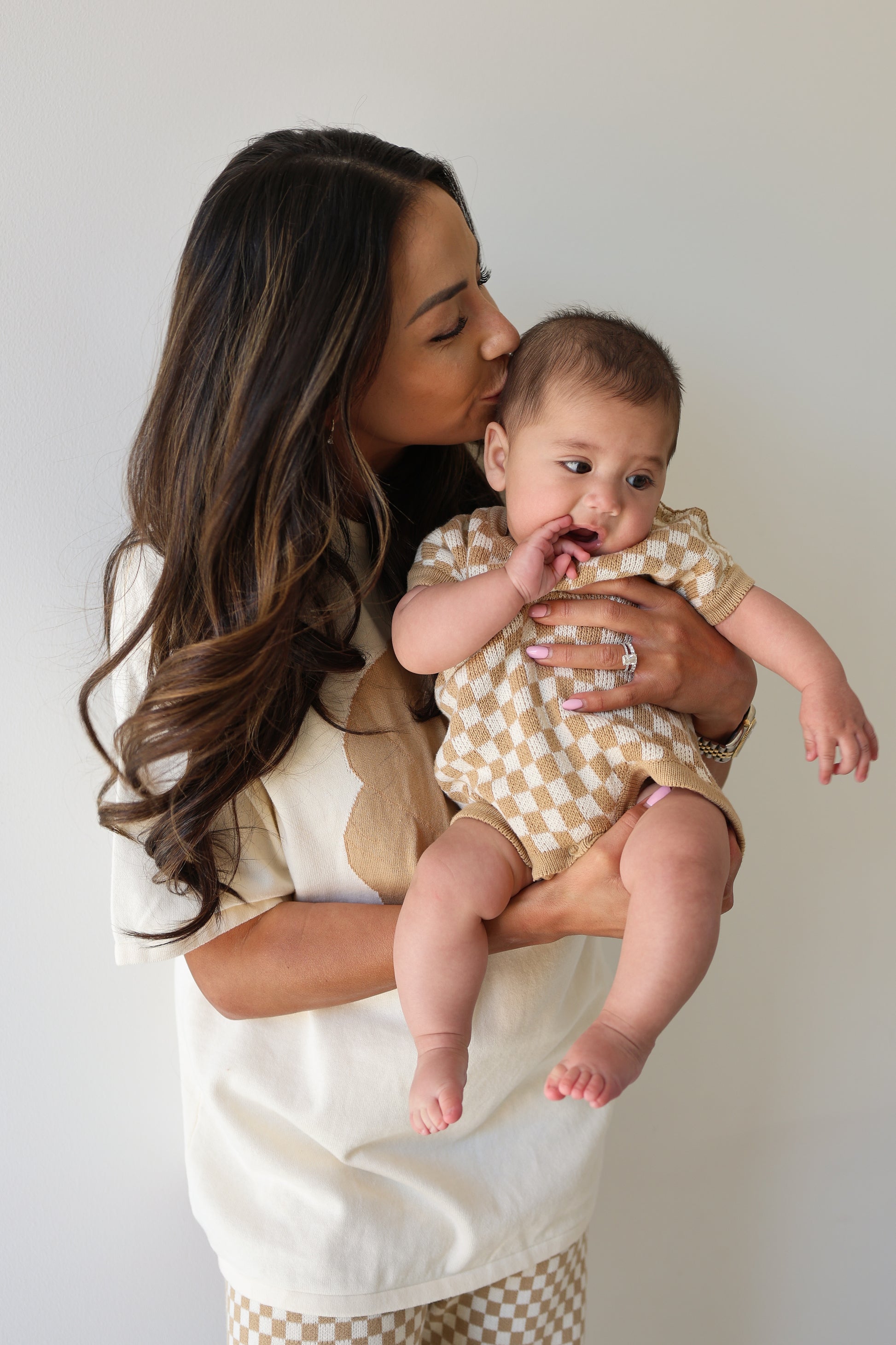 A woman in a cream and beige top with checkered pants kisses a baby wearing the forever french baby Knit Romper in Latte Check, both posed against a plain light background.