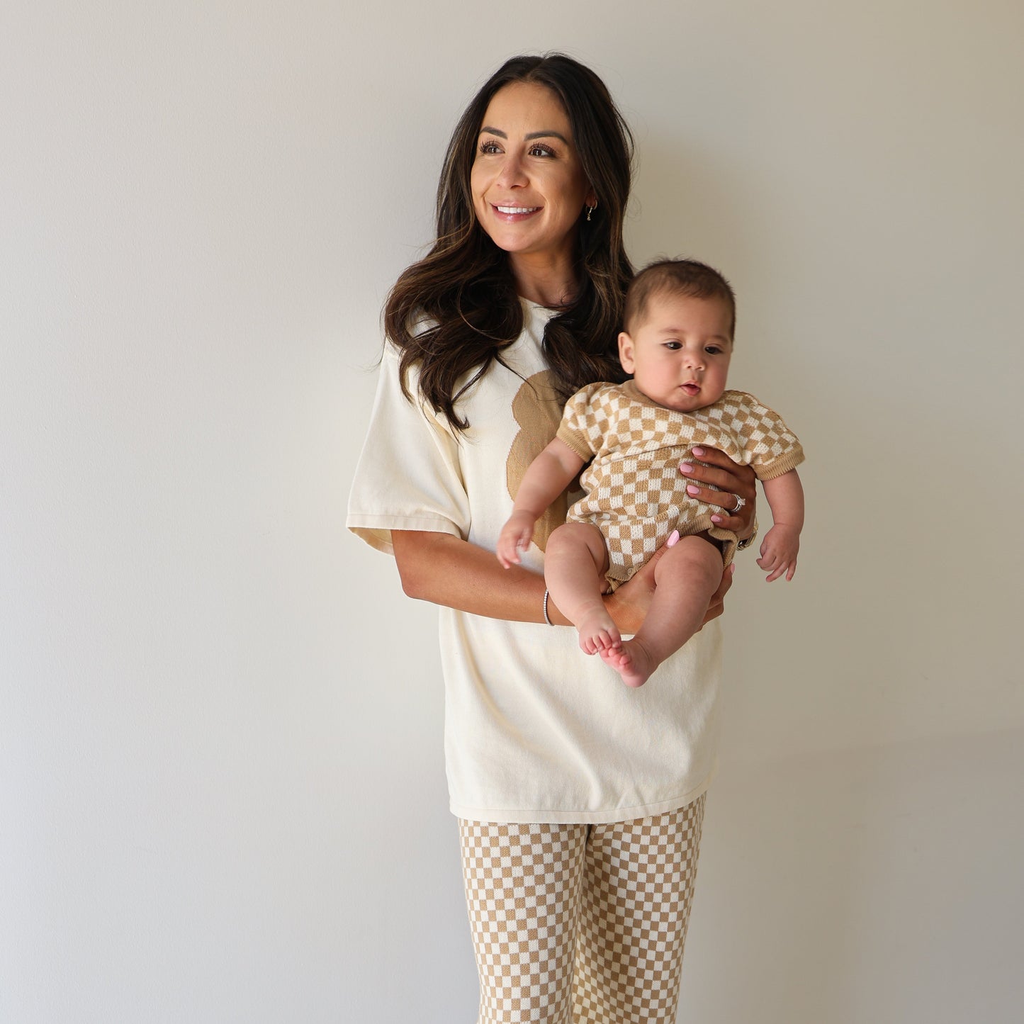 A woman with long dark hair smiles while holding a baby. She wears the forever french baby Women's Knit Pant Set | Croissants, featuring an oversized cream top and beige checkered pants. The baby is in a matching beige and white checkered cotton outfit.