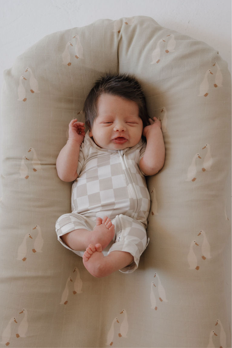 A newborn with dark hair sleeps peacefully on a cushioned surface, wearing the forever french baby Bamboo Zip Shortie | Checkerboard—breathable, light-colored baby clothing. The cushion displays small, simple animal illustrations.