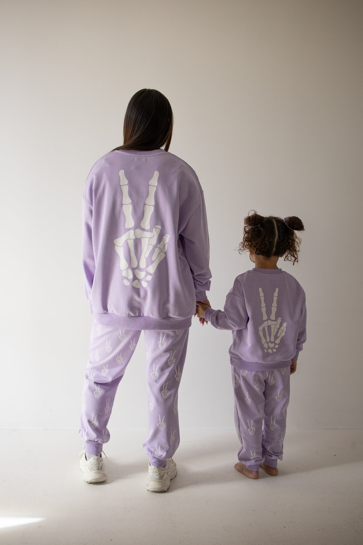 An adult and child, dressed in matching lavender Enchanted Skelly sweat sets from forever french baby—with white skeleton handprints on the backs—stand hand-in-hand, facing away from the camera in a softly lit room, ready for everyday adventures.