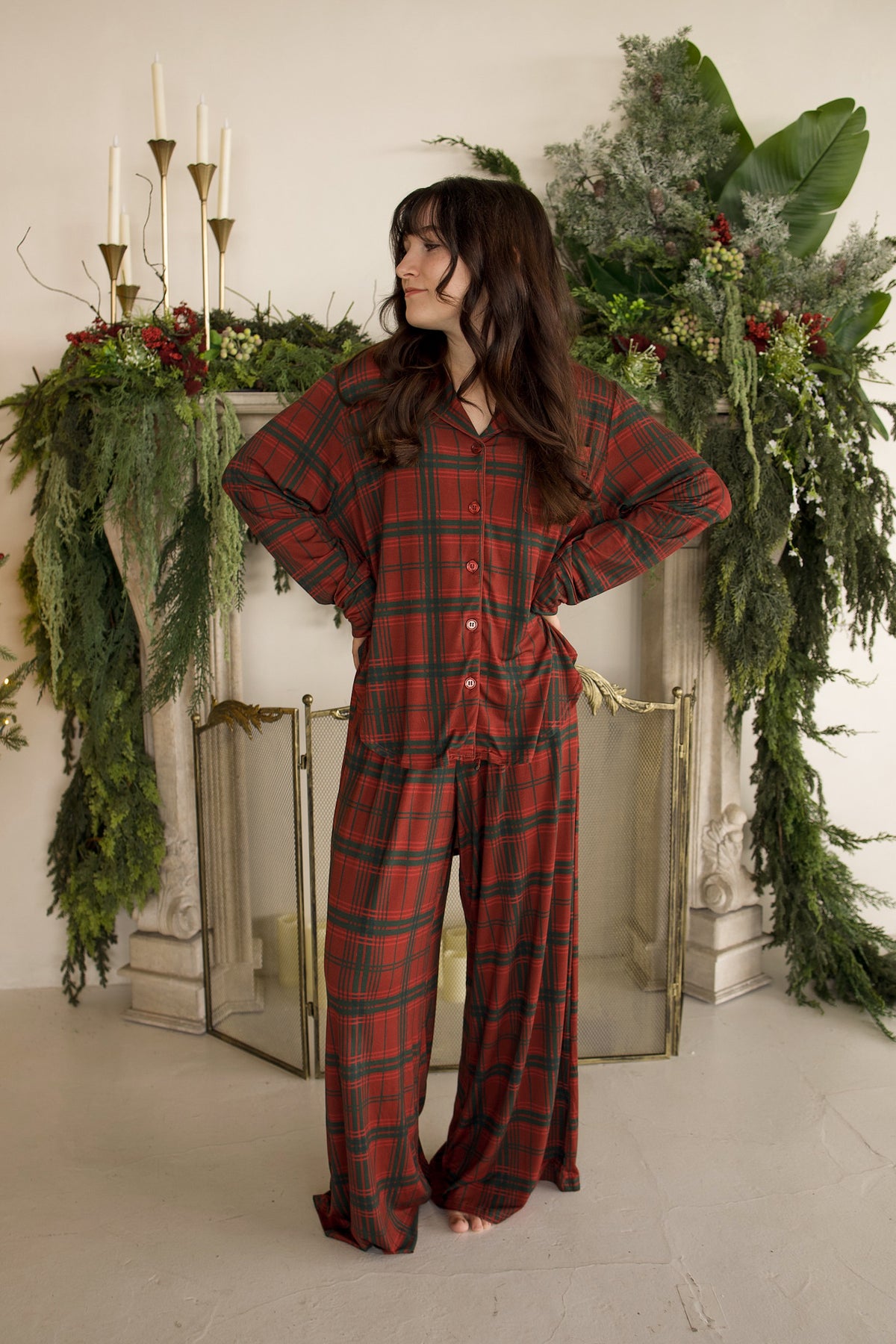 A woman wearing a Women’s Bamboo Button Up Lounge Set in Christmas plaid standing in front of a fireplace decorated with greenery.