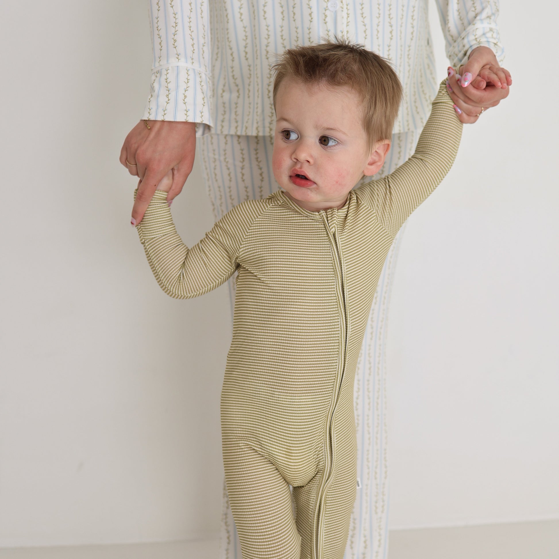 A toddler in a light yellow striped onesie holds the hands of an adult wearing Forever French Baby's Bamboo Zip Pajamas in Willow Stripe Ribbed as they help the child stand and walk.