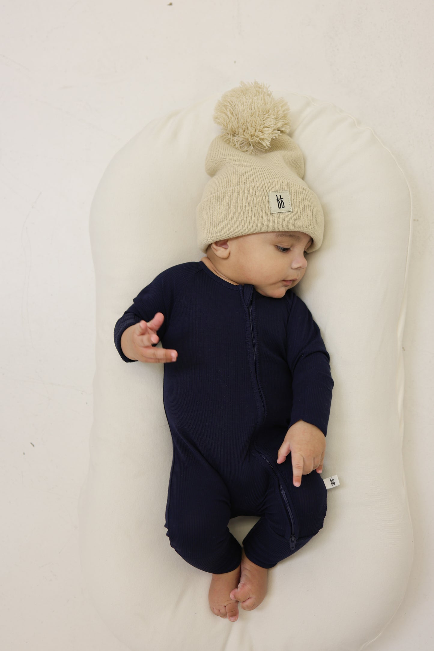 A baby in forever french baby Bamboo Zip Pajamas | Blueberry Ribbed and a beige knit pom-pom hat lies on a white cushioned surface, looking to the side with one hand raised and bare feet showing.