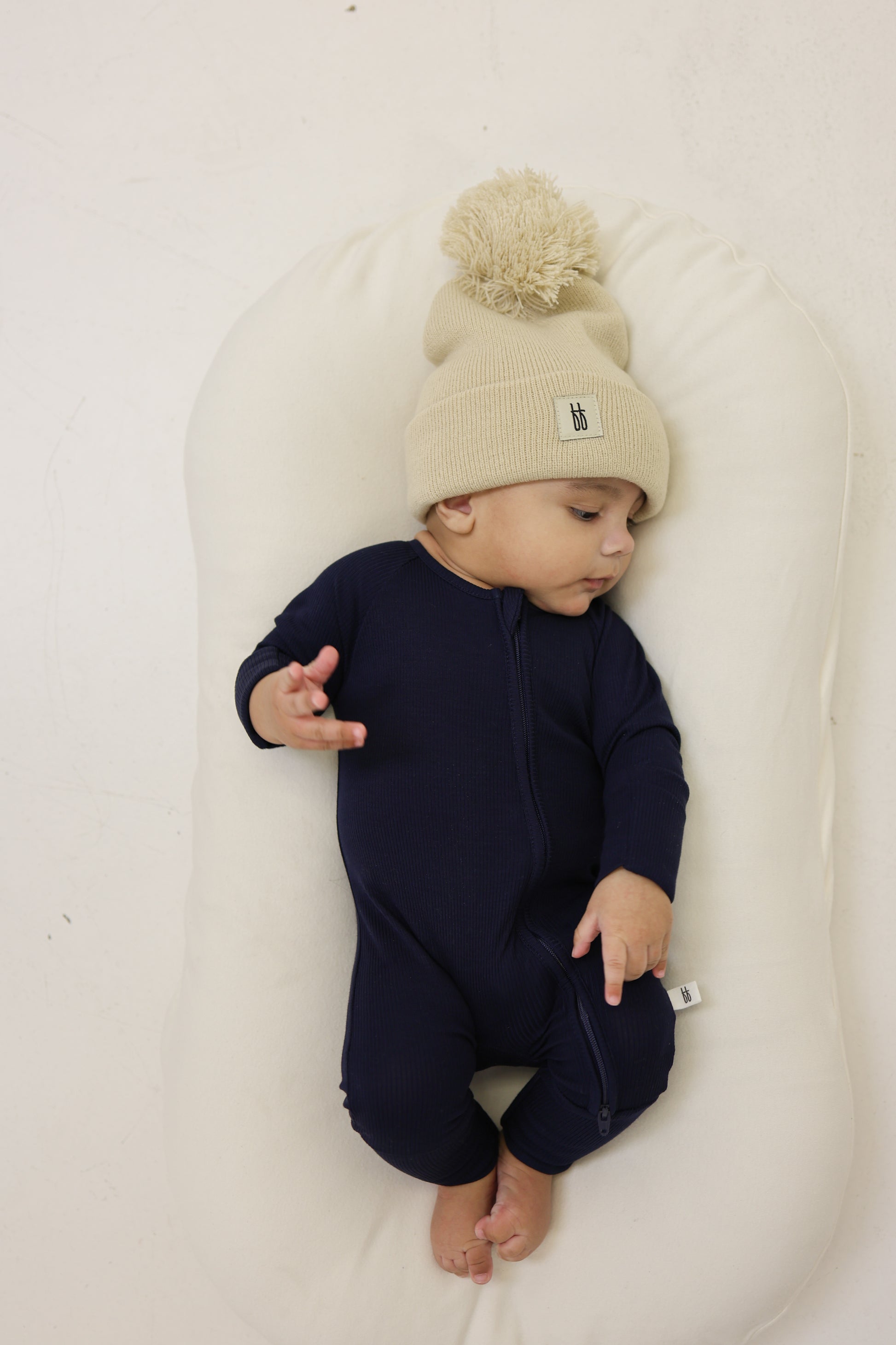A baby in forever french baby Bamboo Zip Pajamas | Blueberry Ribbed and a beige knit pom-pom hat lies on a white cushioned surface, looking to the side with one hand raised and bare feet showing.