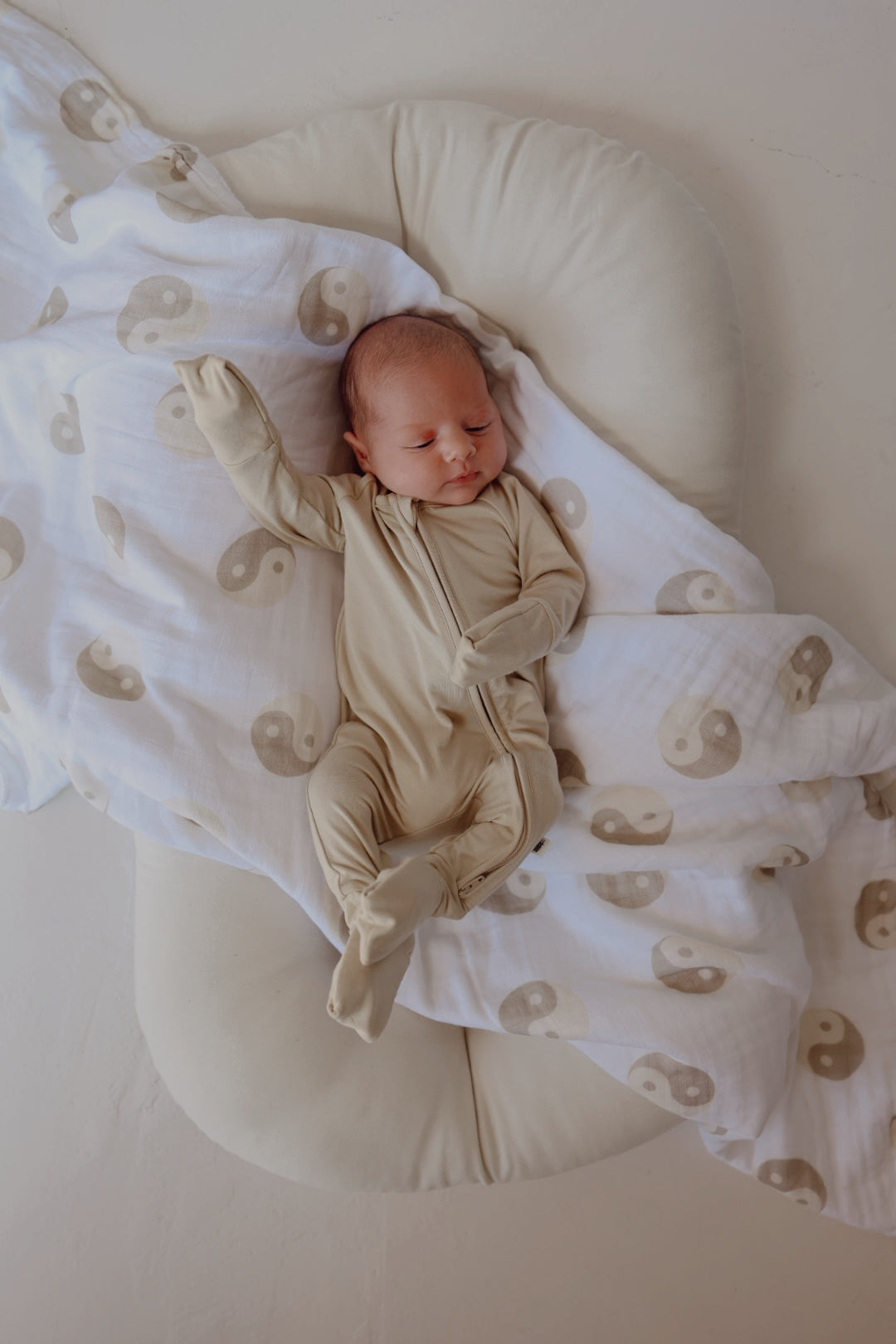 A baby sleeps peacefully on a cushioned surface, wearing the hypo-allergenic Bamboo Zip Pajamas in the Limestone color from forever french baby. Partially covered by a white blanket adorned with beige yin-yang symbols, the scene is serene and minimalistic.