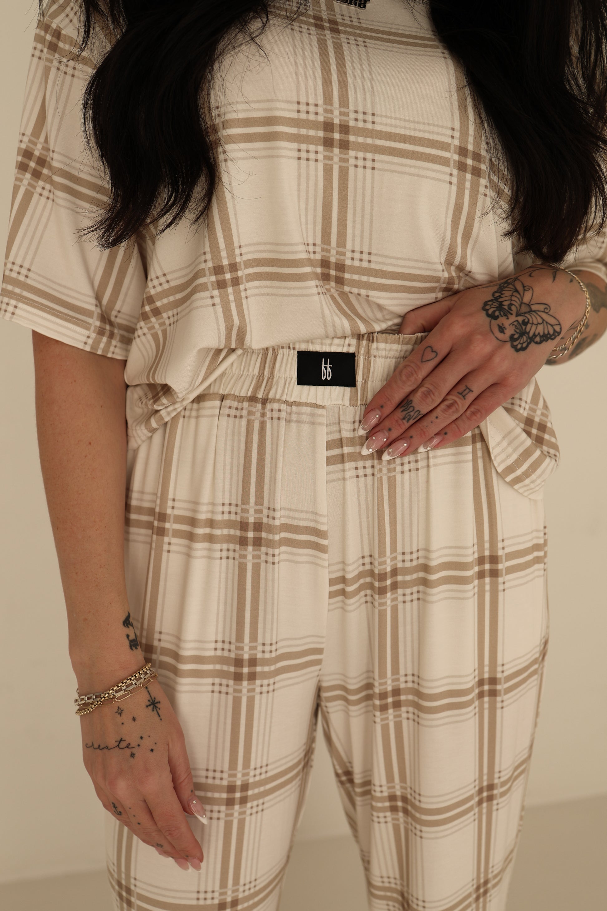 A woman models the Women's Boxy Tee Lounge Set in Ellis Plaid by forever french baby, showing tattoos and a gold bracelet. Only her torso and hands are visible. The hypo-allergenic outfit features a black "bb" tag for style and comfort.