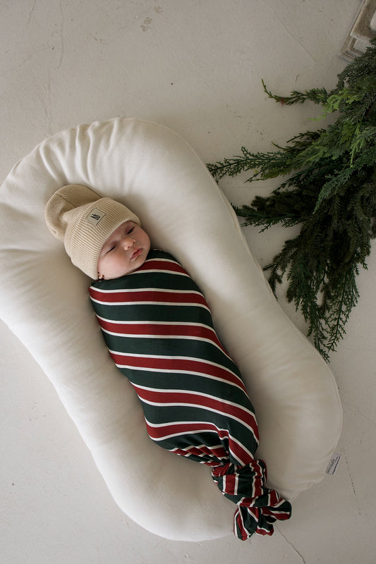 A baby wearing a tan beanie lies in a crescent-shaped lounger, wrapped in a Bamboo Swaddle | Holiday Stripe.