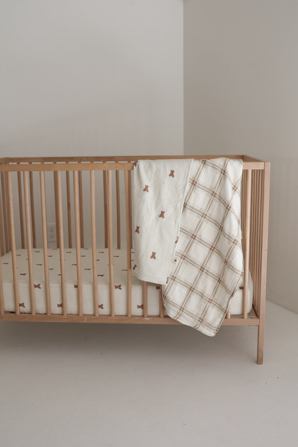 A minimalist wooden crib with a white mattress features two forever french baby Muslin Quilts in the Teddy & Ellis Plaid draped over the side, set against a plain white wall and floor.