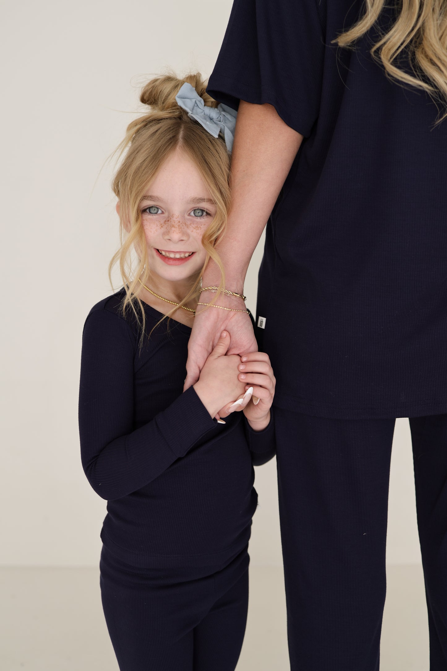 A young girl with blonde hair in a blue scrunchie smiles while holding hands with an adult, both dressed in matching forever french baby Bamboo Two Piece Pajamas | Blueberry Ribbed. The adult is partially out of frame.
