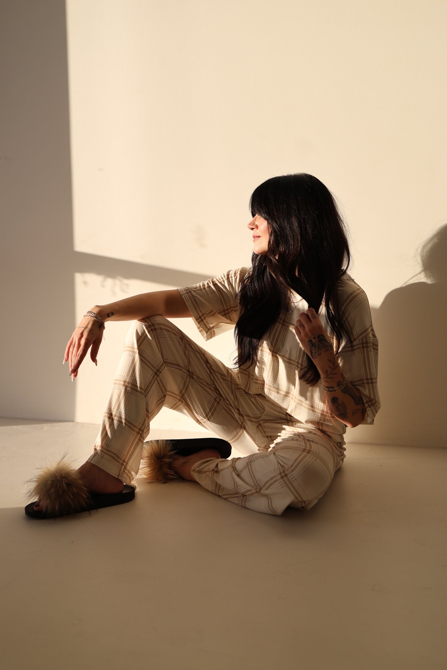 A woman with long dark hair sits on the floor in sunlight, wearing the forever french baby Women's Boxy Tee Lounge Set in Ellis Plaid. She looks to the side, one arm resting on her knee as soft shadows fall behind her.