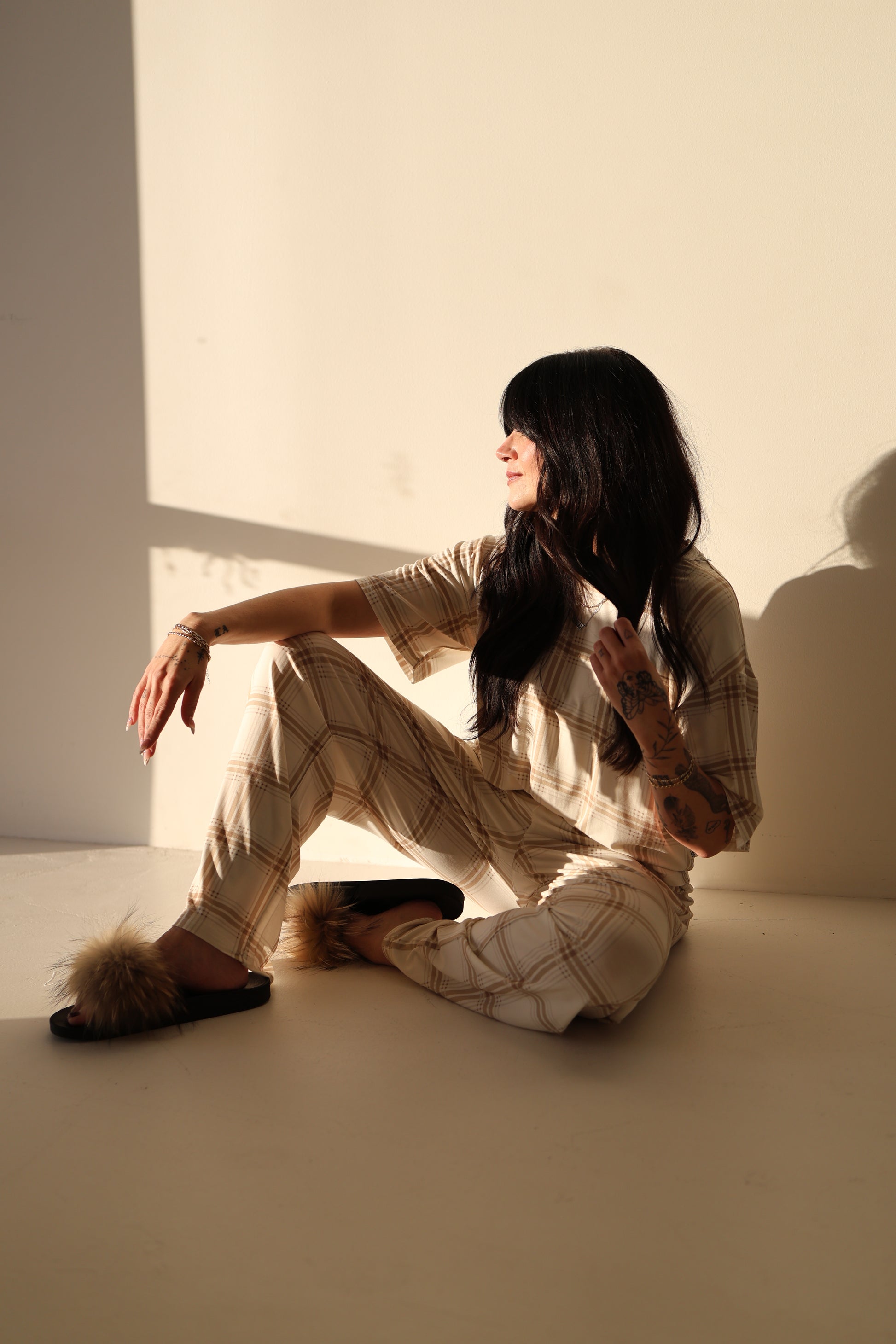 A woman with long dark hair sits on the floor in sunlight, wearing the forever french baby Women's Boxy Tee Lounge Set in Ellis Plaid. She looks to the side, one arm resting on her knee as soft shadows fall behind her.