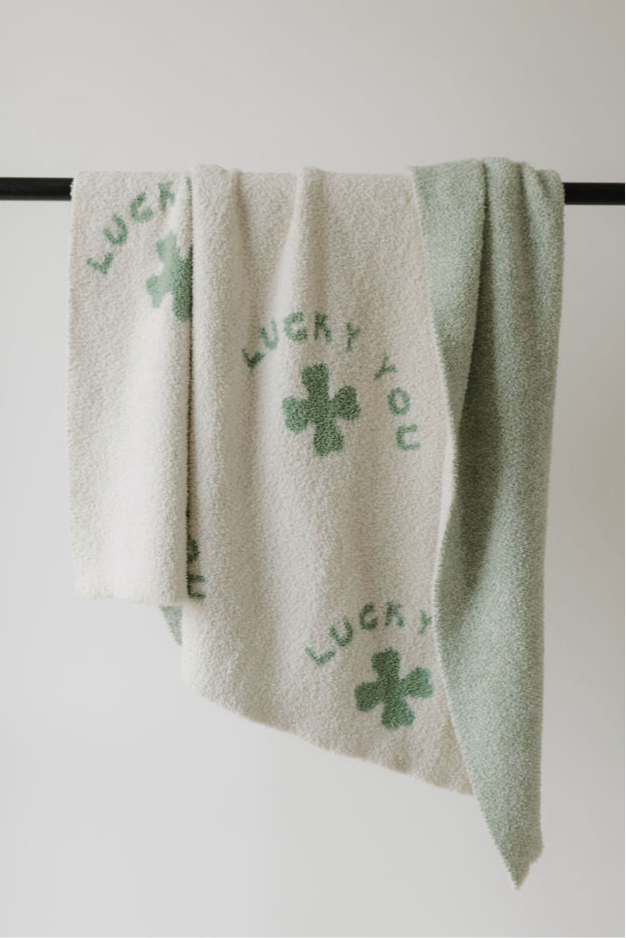 The Plush Blanket | Lucky You by forever french baby is a soft OEKO Standard 100 certified microfiber towel with a green side and a white side featuring green clovers and “LUCKY YOU,” shown draped over a black rod on a white background.