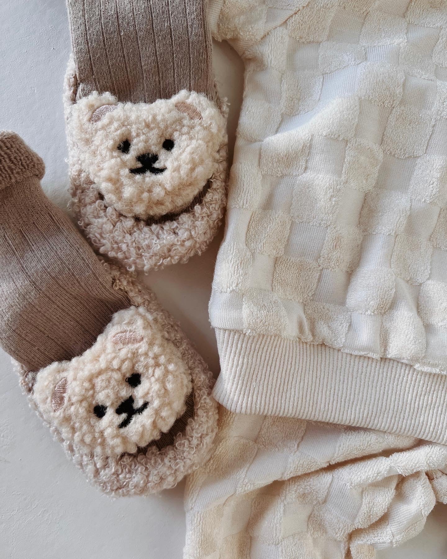 Close-up of a cozy outfit featuring beige and white checkered fluffy pajamas, complemented by the adorable Teddy Bear Slipper Socks from forever french baby. The light brown knitted slipper socks feature bear faces made of a soft, wool-like material, adding an adorable detail to the ensemble. The background is a plain white.