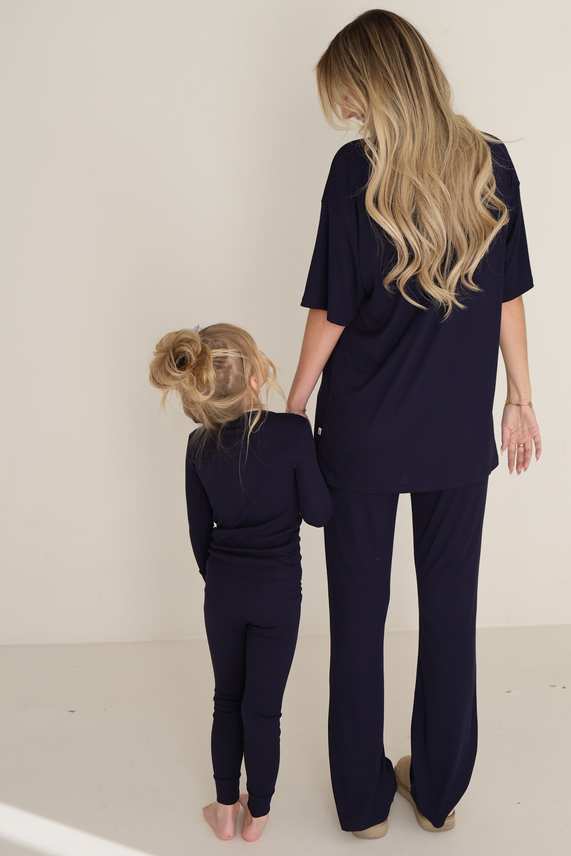 A woman and a young girl stand barefoot, holding hands in matching blueberry ribbed bamboo lounge sets by forever french baby, both facing away from the camera against a light background.