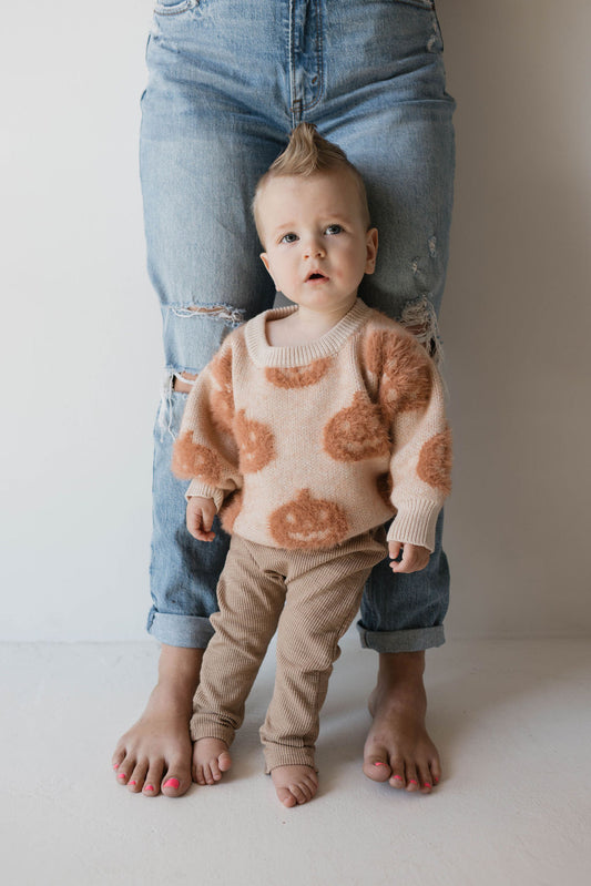 A toddler stands barefoot in front of an adult's legs, leaning back slightly into them. The toddler wears a Cozy Sweater | Pumpkin from forever french baby with brown fuzzy circles, along with brown corduroy pants, and has a small mohawk hairstyle. The adult is barefoot and dressed in ripped jeans.