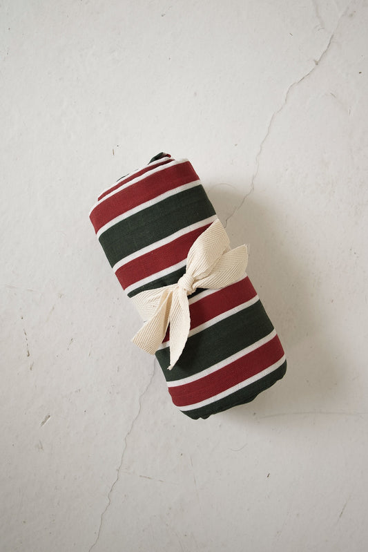 A rolled Muslin Swaddle | Holiday Stripe in dark green, red, and white holiday stripes, tied with a beige ribbon.