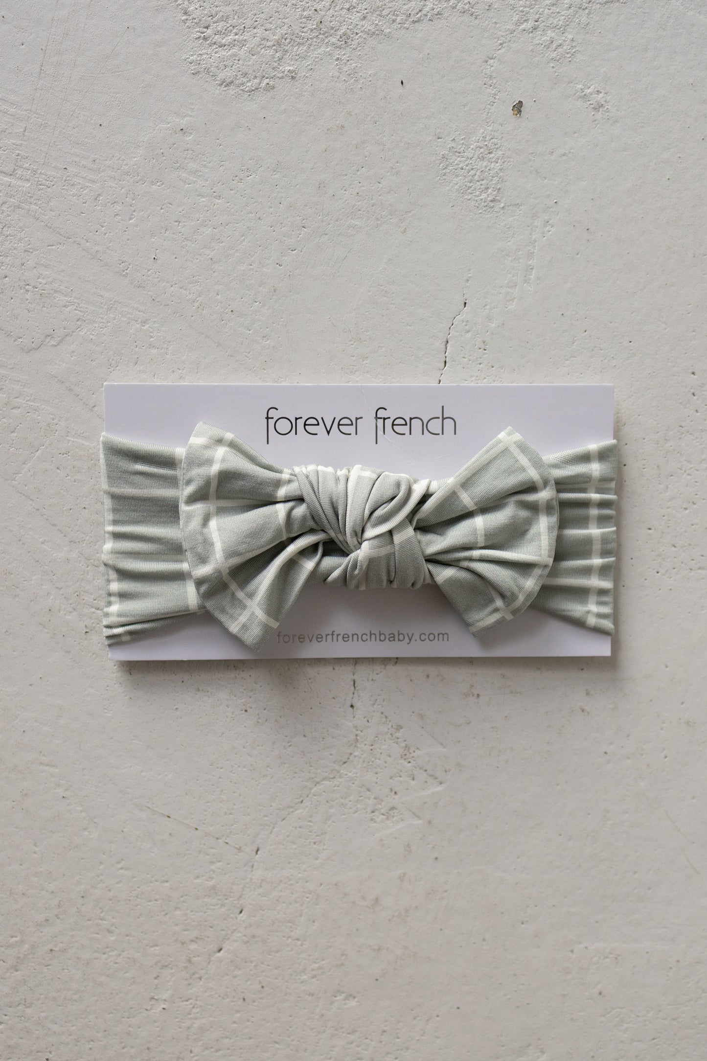 A Bamboo Head Wrap in the “Foggy Grid” design—a sage green plaid baby headband with a large bow, made from ultra-soft bamboo and fully adjustable—is showcased on a white forever french baby branded card against a textured light gray background.