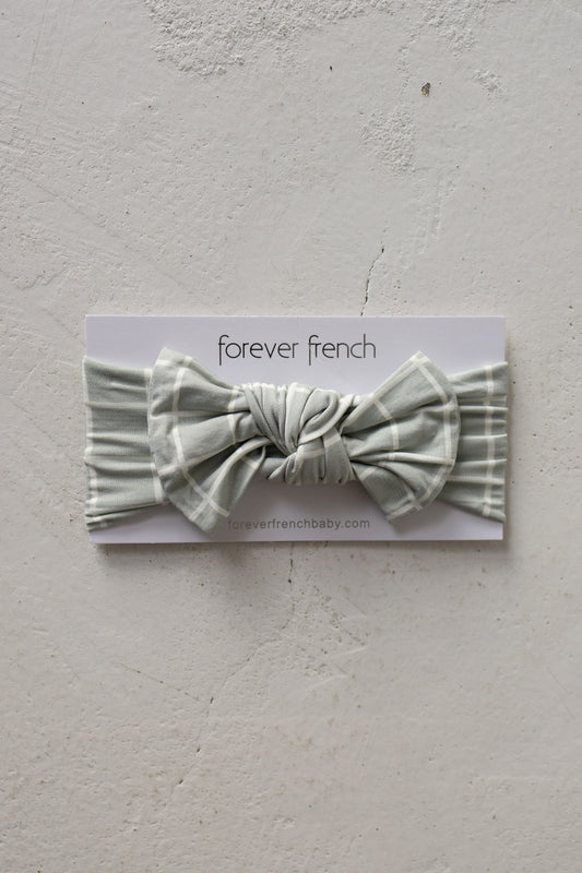 A Bamboo Head Wrap in the “Foggy Grid” design—a sage green plaid baby headband with a large bow, made from ultra-soft bamboo and fully adjustable—is showcased on a white forever french baby branded card against a textured light gray background.
