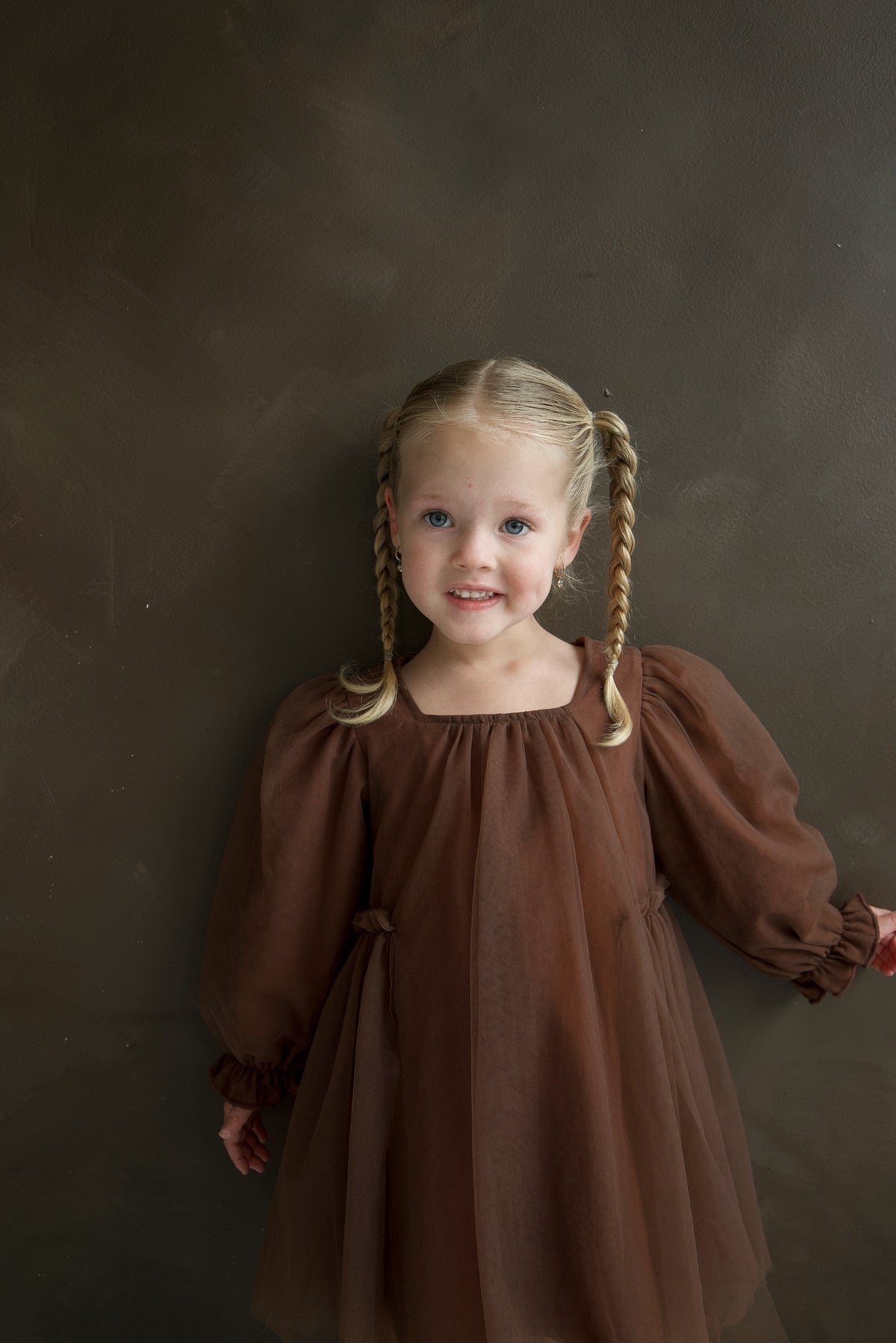 A young girl wearing a Long Sleeve Tutu Dress | Cocoa, a long-sleeved brown festive tutu dress with bow detail.