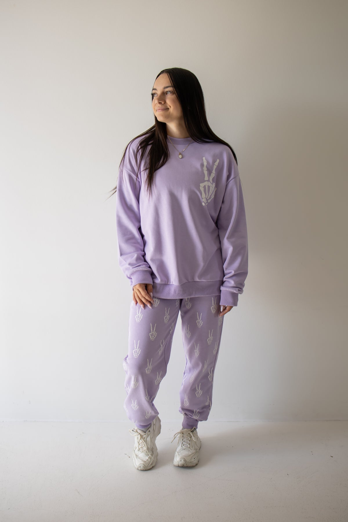 A woman stands against a plain wall wearing the Women’s Sweat Set from forever french baby’s Enchanted Skelly collection. This matching lavender set features white patterns and is paired with white sneakers. She smiles and gazes off to the side, making this effortlessly stylish outfit a perfect pick for your next spooky season look.