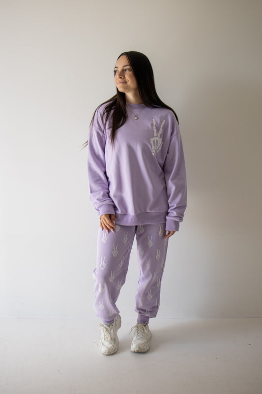 A woman stands against a plain wall wearing the Women’s Sweat Set from forever french baby’s Enchanted Skelly collection. This matching lavender set features white patterns and is paired with white sneakers. She smiles and gazes off to the side, making this effortlessly stylish outfit a perfect pick for your next spooky season look.