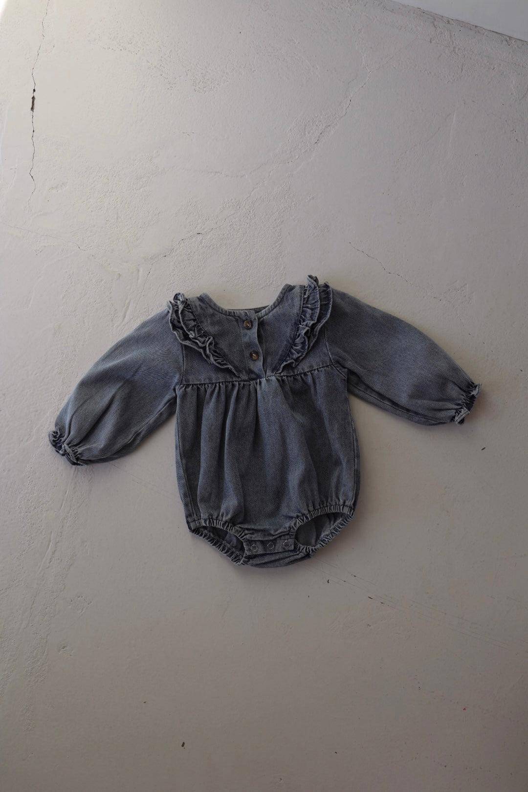 The forever french baby Denim Romper | Ruffle is a long-sleeve, tunic-style denim romper for babies with a ruffled collar and front buttons, shown laid flat on a light-colored textured surface.