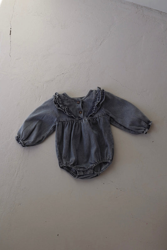 The forever french baby Denim Romper | Ruffle is a long-sleeve, tunic-style denim romper for babies with a ruffled collar and front buttons, shown laid flat on a light-colored textured surface.