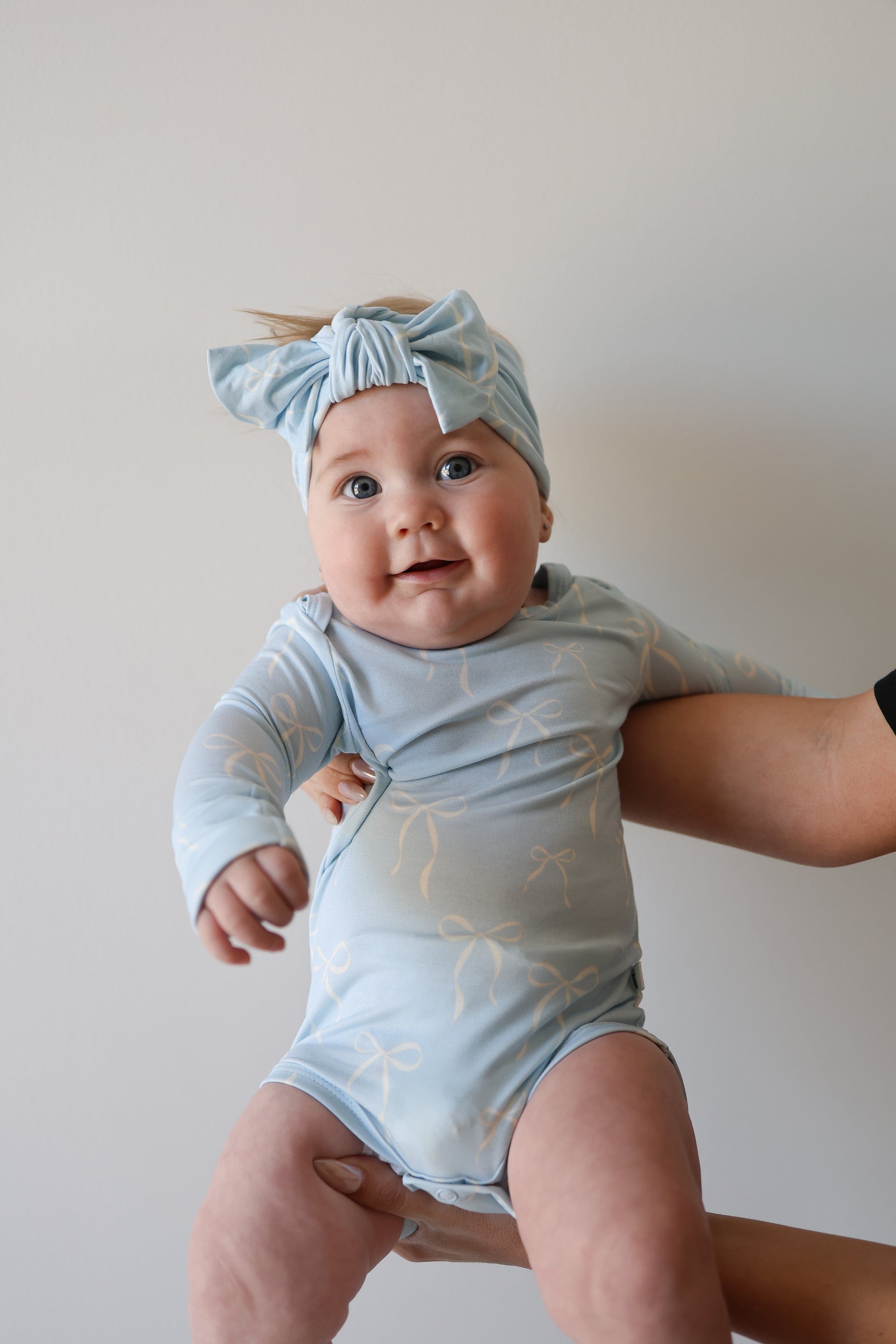 A smiling baby wears the forever french baby Long Sleeve Bamboo Snapsuit | Blue Bows with a matching headband, held up by an adult’s hand against a white background, highlighting this soft, hypoallergenic bamboo outfit.