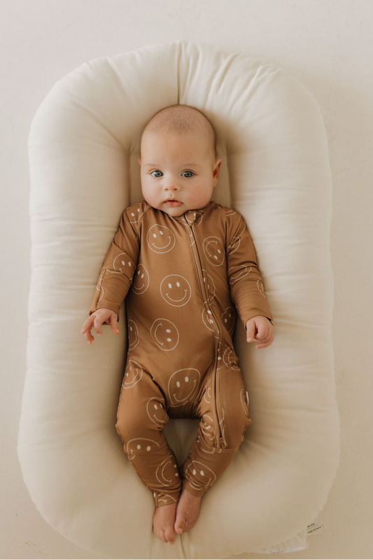 A baby in forever french baby Bamboo Zip Pajamas | Sandstone Smiles, a hypo-allergenic brown onesie with white smiley faces, lies calmly on a soft white lounger.