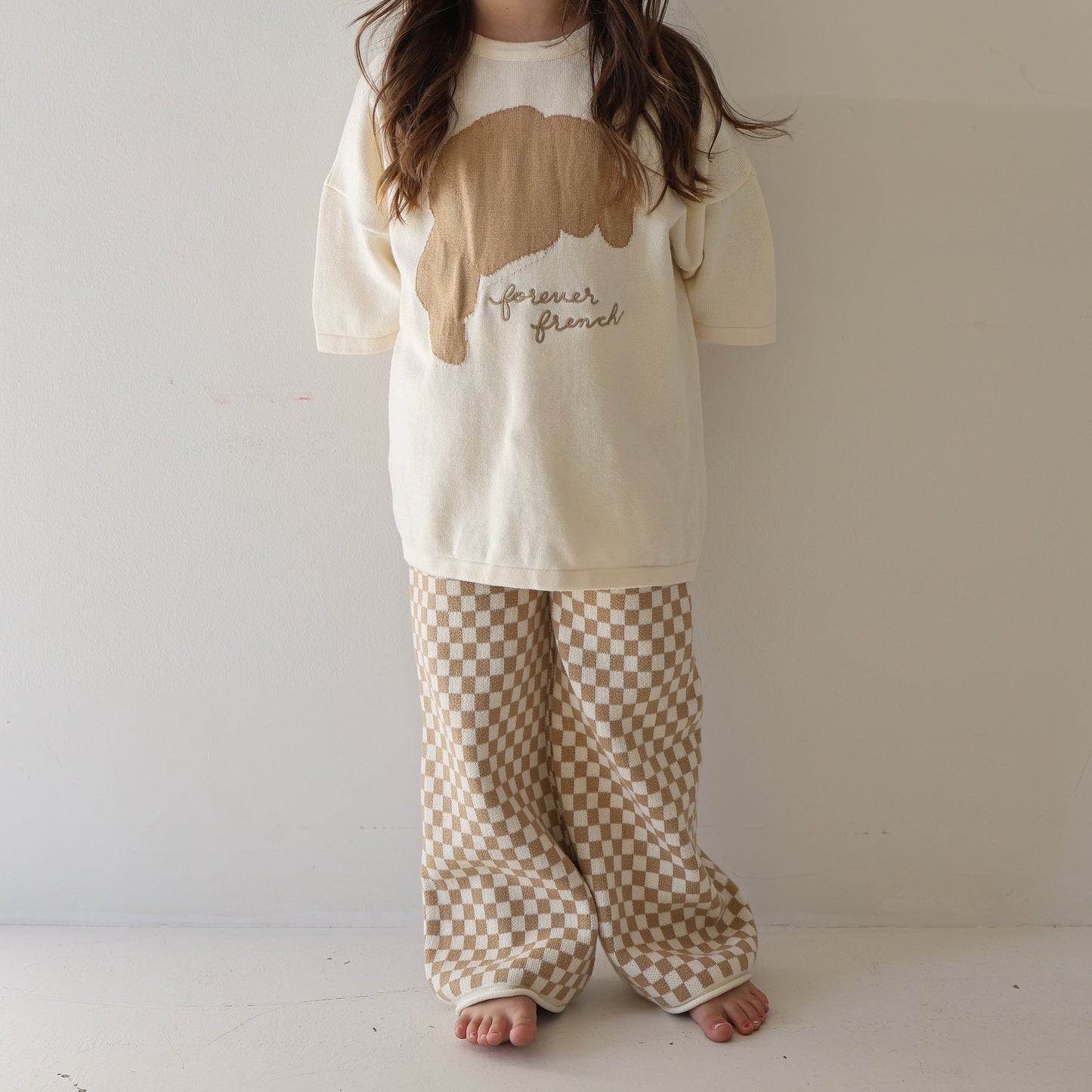 A child stands barefoot against a plain wall, wearing the forever french baby Knit Pant Set | Croissants—an oversized cream top with "forever brunch" graphic and loose beige-and-white checkered pants; long brown hair partially covers their face.