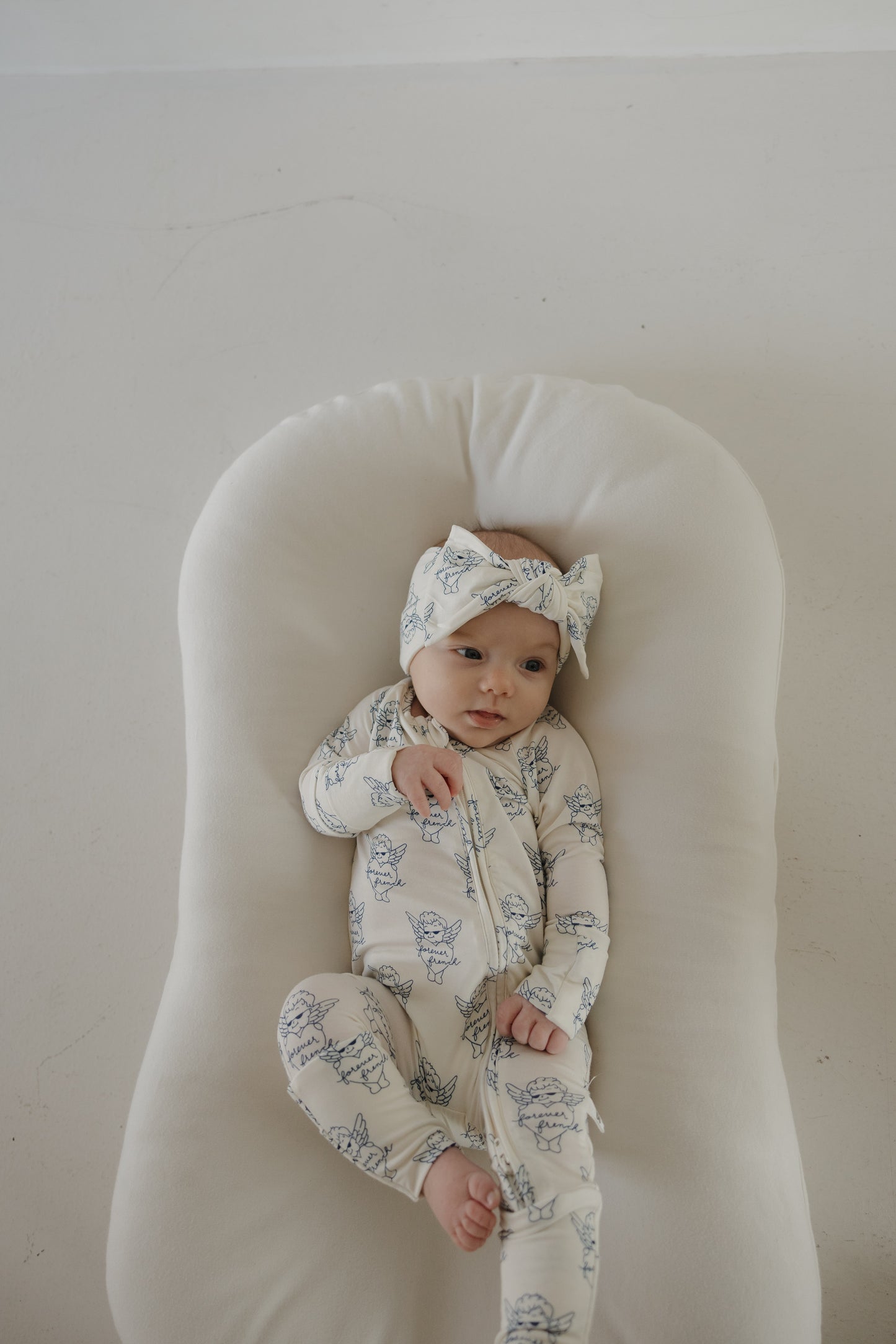 A baby in forever french baby's Bamboo Zip Pajamas by ff Los Angeles—a matching white hat and onesie with blue animal prints—lies on a soft cushion, gazing gently to the side.
