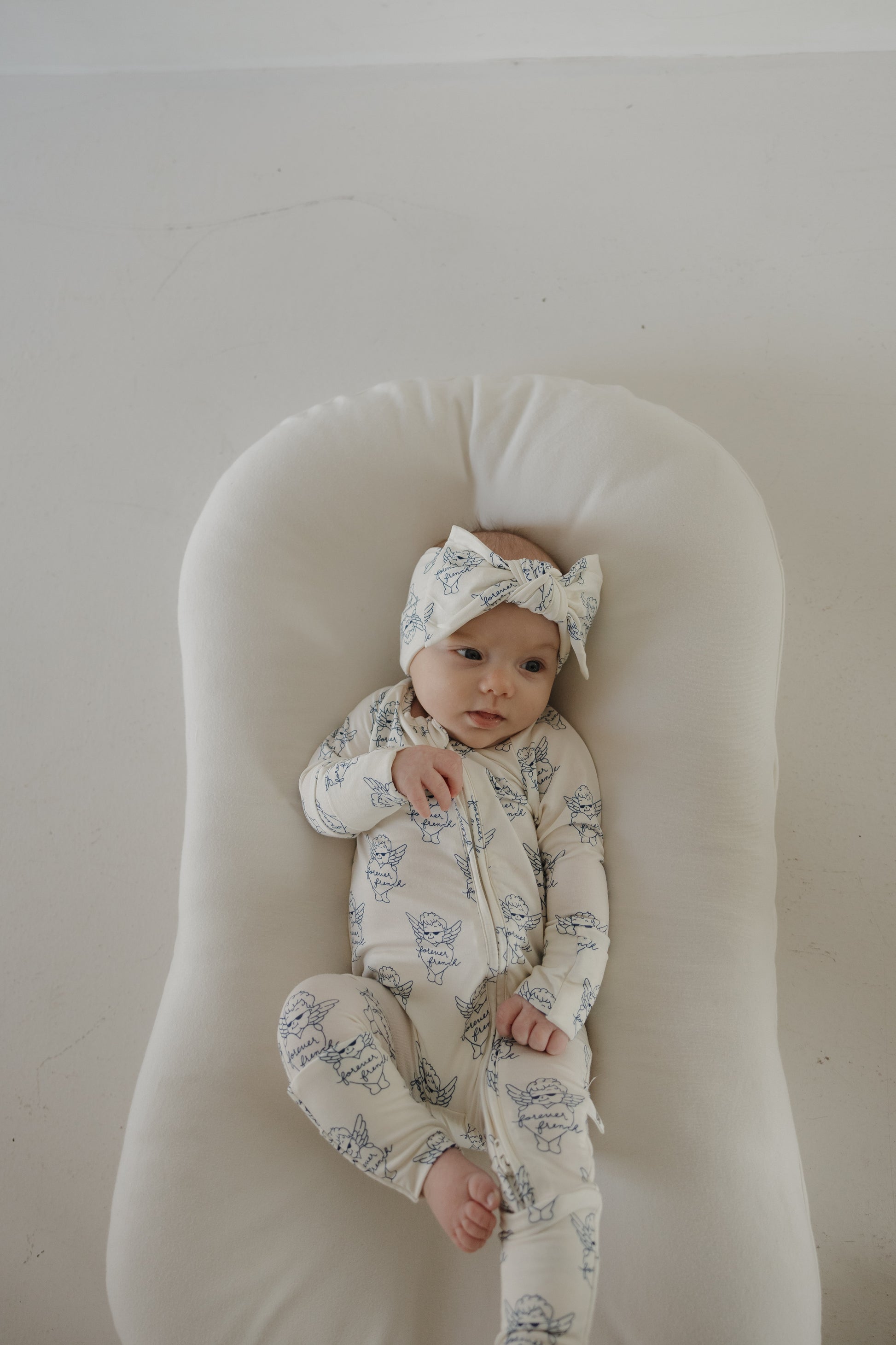 A baby in forever french baby's Bamboo Zip Pajamas by ff Los Angeles—a matching white hat and onesie with blue animal prints—lies on a soft cushion, gazing gently to the side.