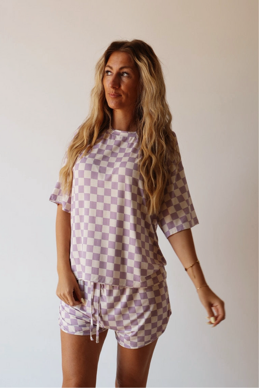 A woman with long wavy hair wears the forever french baby Women's Bamboo Short Set in Grape Soda, featuring lavender and white checks, against a plain light background.
